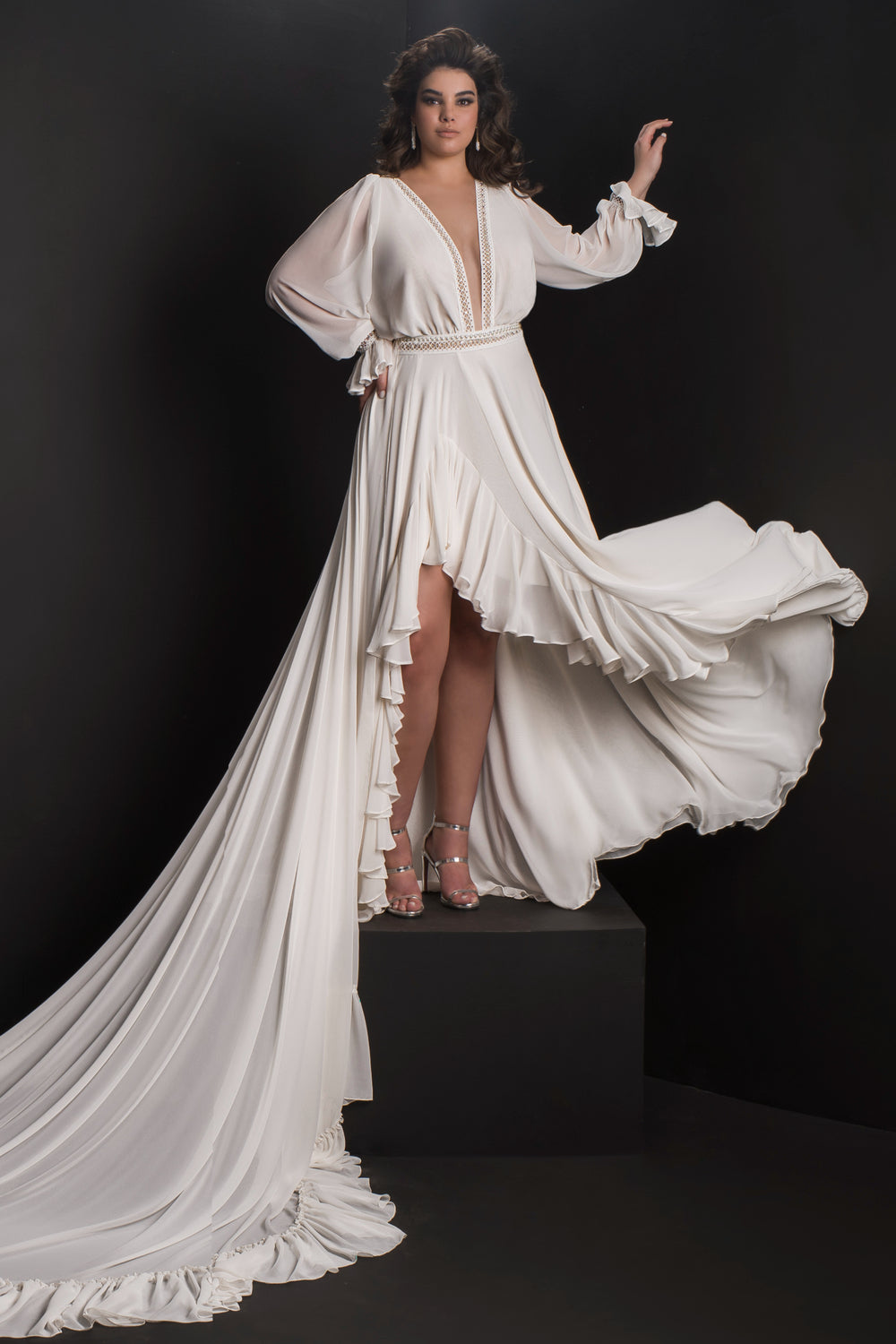 אישה פוזלת על במה שחורה לבושה בשמלת תרזה מקולקציית Bridal Plus 2019 של דרור קונטנטו, עם מחשוף V עמוק, שרוולים ארוכים, גימור קפלים ושובל דרמטי — מושלמת לחתונות אלגנטיות במידות גדולות.