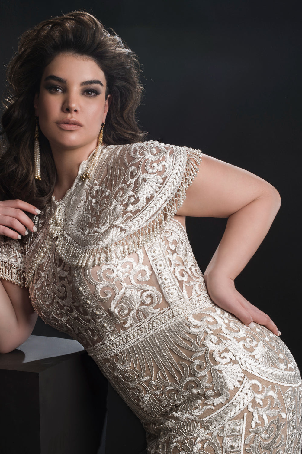 אישה עם שיער כהה ושופע לובשת את שמלת הכלה "Bridal Plus Collection 2019 - Amelie" בצבע שמנת, מעוטרת ברקמה מורכבת, מבית Dror Kontento, בשילוב עם עגילים בולטים, ומצטלמת על רקע כהה.