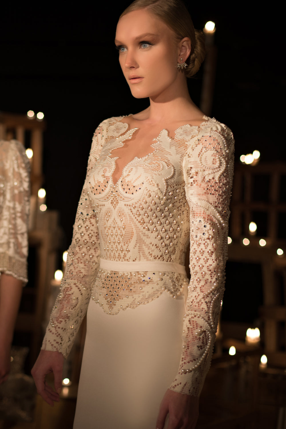 אישה לבושה בשמלת תחרה לבנה אלגנטית עם שרוולים ארוכים, עיטורי חרוזים מורכבים ומותניים צמודות, עומדת בתאורה רכה ועמומה עם נרות מטושטשים, ומציגה את קולקציית הכלות 2015 - 05.