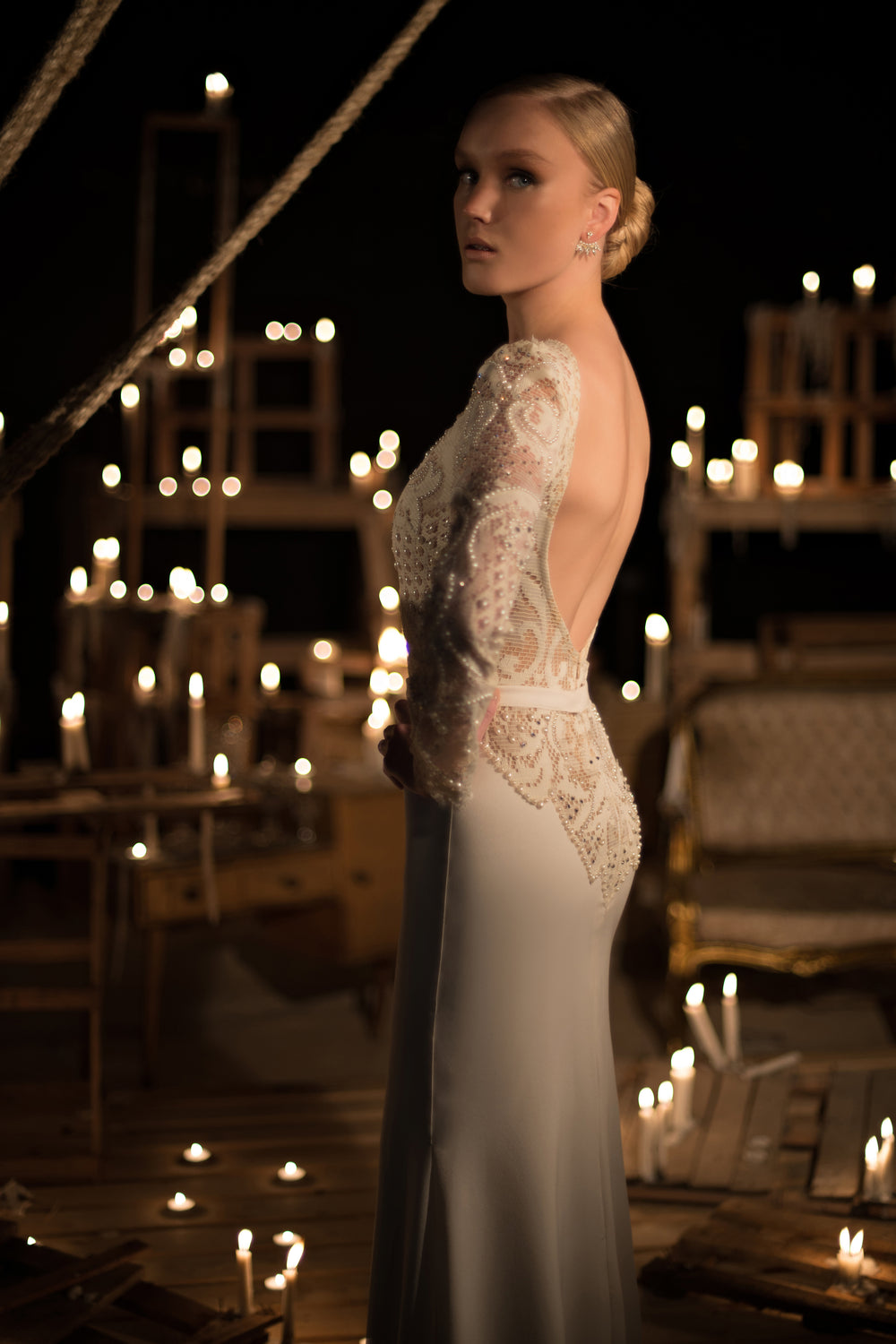 אישה בשמלת כלה לבנה עם שרוולים ארוכים וגב חשוף מקולקציית הכלות 2015 - 05 עומדת בין נרות בוערים על משטחי עץ, ומביטה מעבר לכתפה. הרקע הכהה יוצר אווירה דרמטית ואלגנטית.