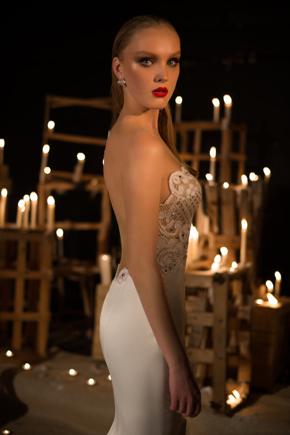 אישה מקולקציית הכלות 2015 - שמלת כלה בעיצוב אישי עם 12 רצועות ללא כתפיות ועיטורי חרוזים מורכבים עומדת בפרופיל בין ארגזי עץ מוארים בנרות וחול, שיערה החלק ושפתיה האדומות הבולטות יוצרים אווירה דרמטית ונטולת זמן.