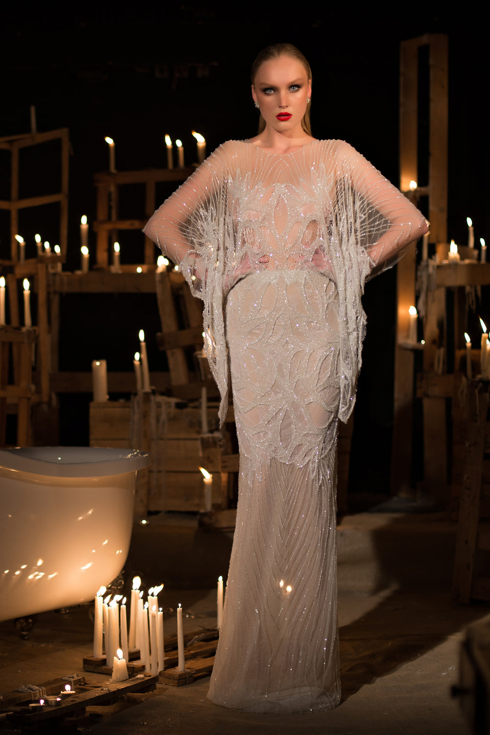 אישה בשמלת "קולקציית כלות 2015 - 13" עומדת בביטחון, ידיה על מותניה, מוקפת בנרות דולקים על ארגזי עץ, בסביבה דרמטית ומוארת באור עמום.