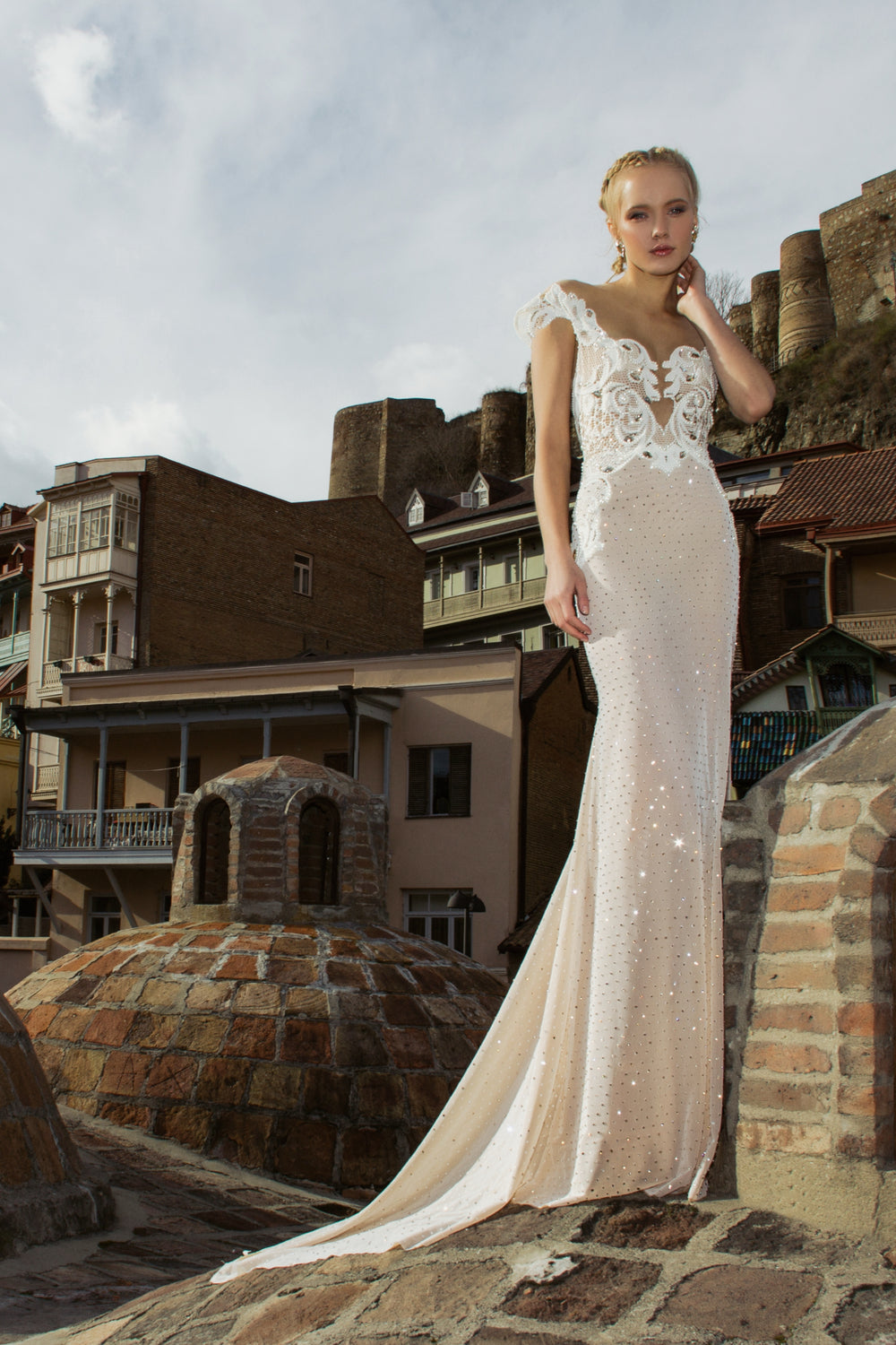 אישה לובשת את קולקציית הכלות 2016 - 02, שמלת כלה לבנה צמודה, בעודה עומדת בחוץ על כיפות לבנים, עם מבנים מסורתיים ומבצר אבן היסטורי ברקע.