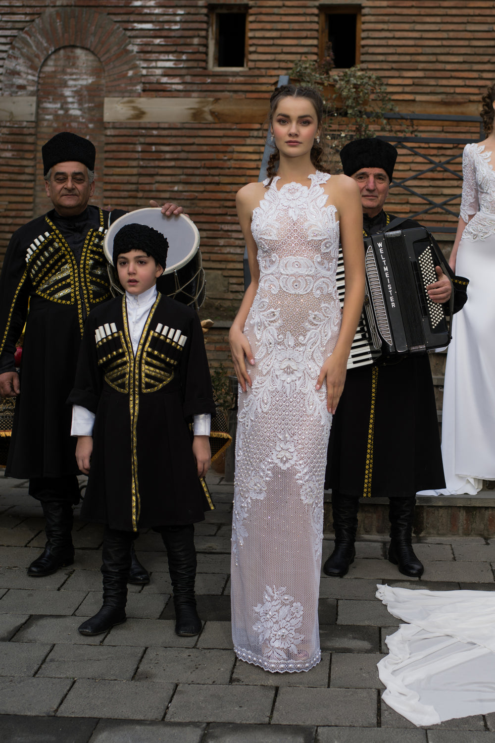 אישה בשמלת תחרה לבנה מקולקציית הכלות 2016 - 08 עומדת לצד נער לבוש בבגד גאורגי מסורתי שחור, בעוד גברים לבושים בבגדים דומים מנגנים על כלי נגינה לפני קיר לבנים.