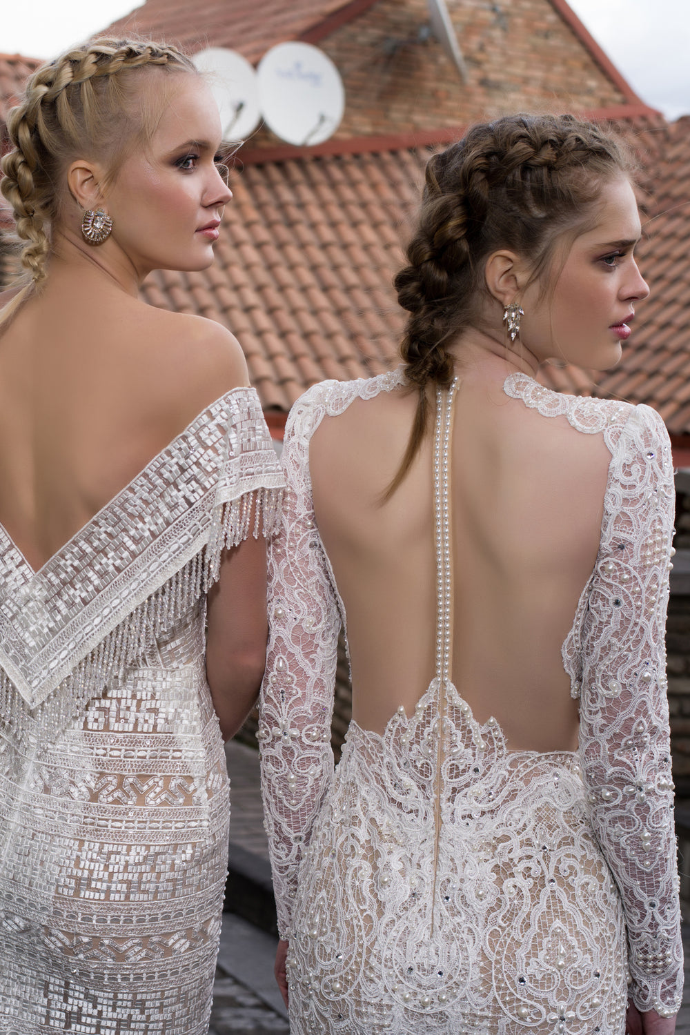 שתי נשים עם צמות בשיערן לובשות שמלות כלה בעיצוב אישי מהקולקציה Bridal Collection 2016 - 05, עומדות בחוץ על גג רעפים וליד קיר לבנים, כשברקע צלחות לווין.