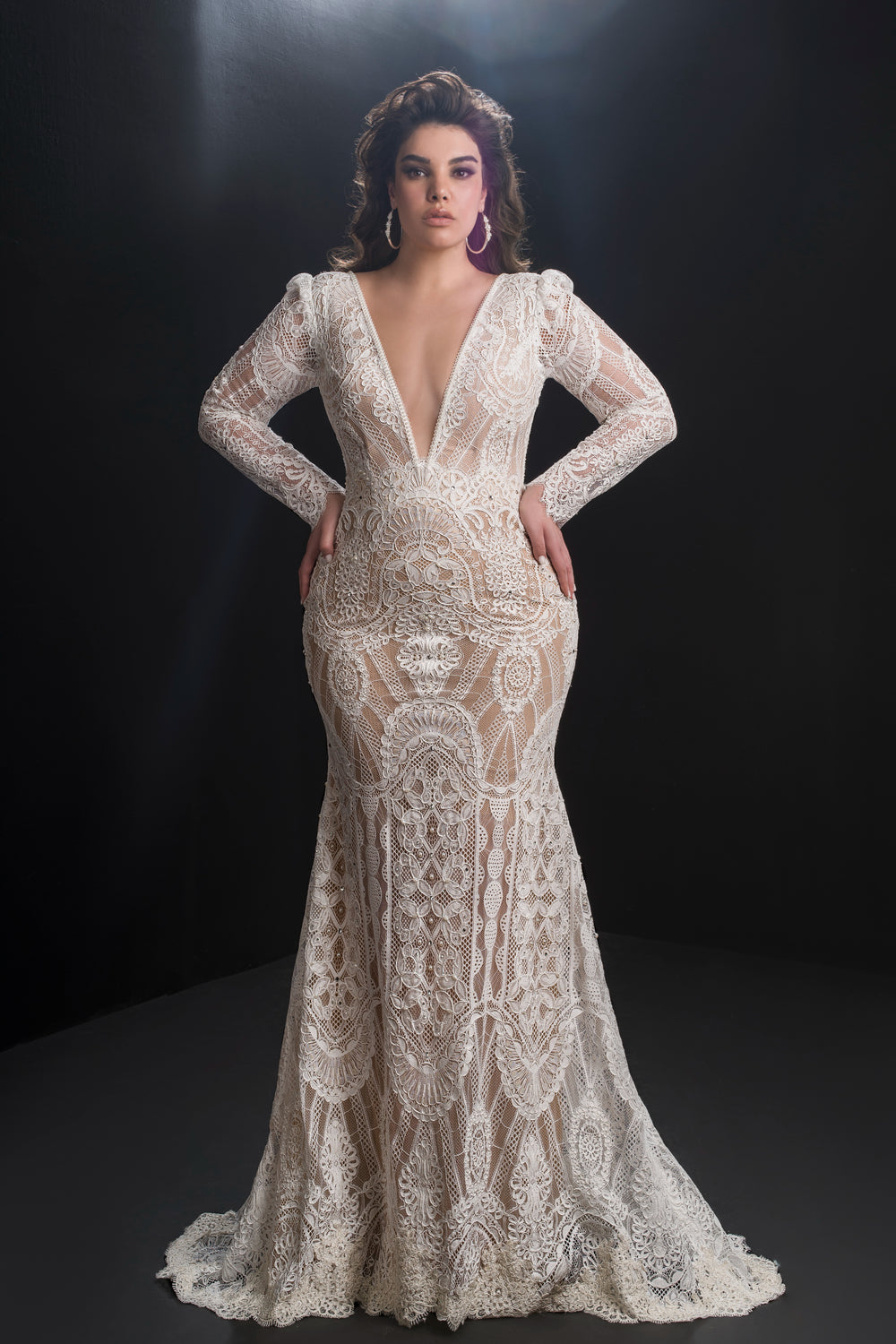 אישה עומדת בביטחון בשמלת Rebeca מקולקציית Bridal Plus 2019 - שמלת תחרה לבנה צמודה עם שרוולים ארוכים, מחשוף V עמוק ודוגמאות מורכבות, על רקע כהה.