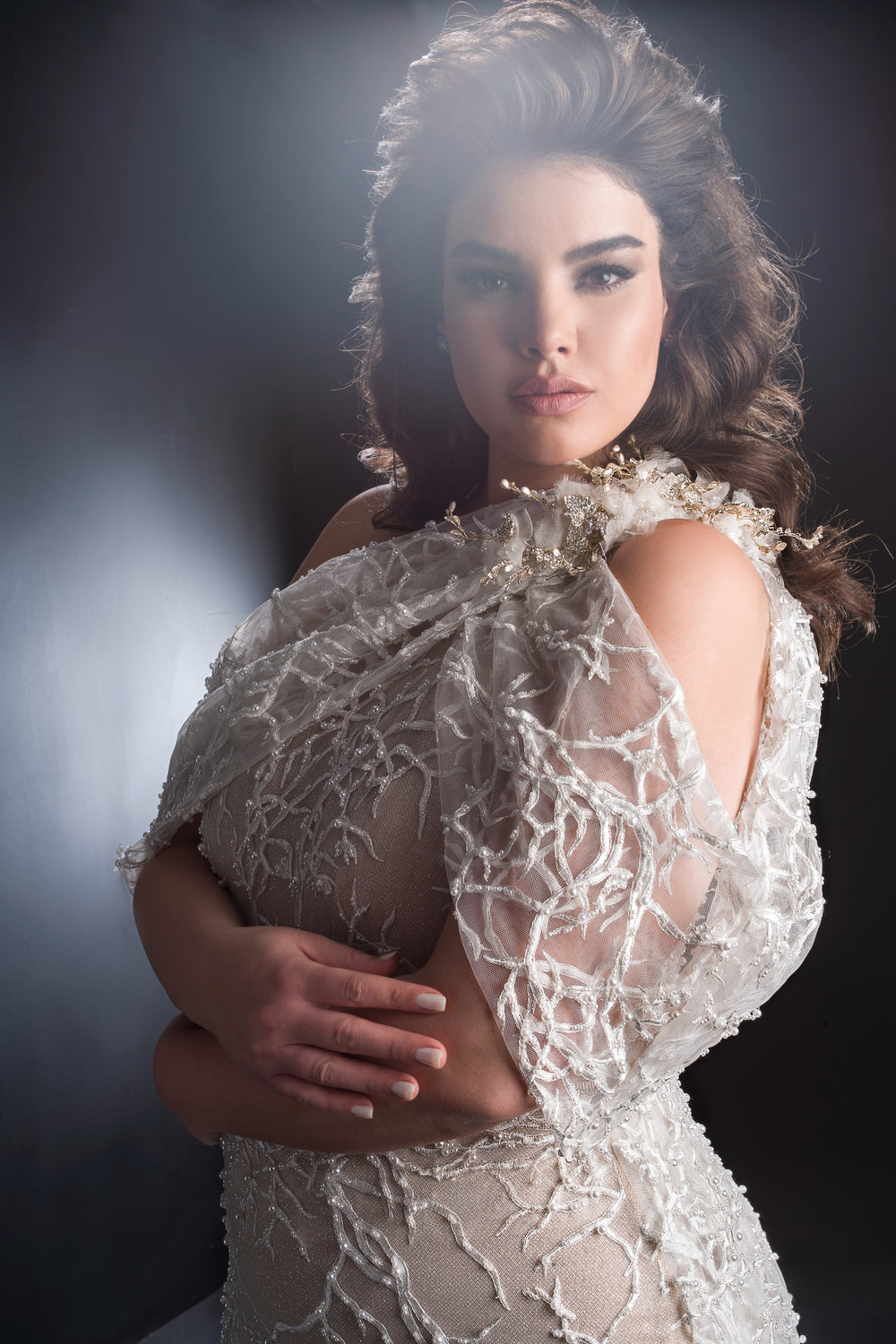 אישה עם שיער ארוך, מלא ונפוח, לובשת שמלת כלה אלגנטית, שקופה ומרוקמת מקולקציית Bridal Plus 2019 - Royal, עומדת עם זרועות שלובות ומביטה בביטחון למצלמה על רקע כהה ומואר באור רך.