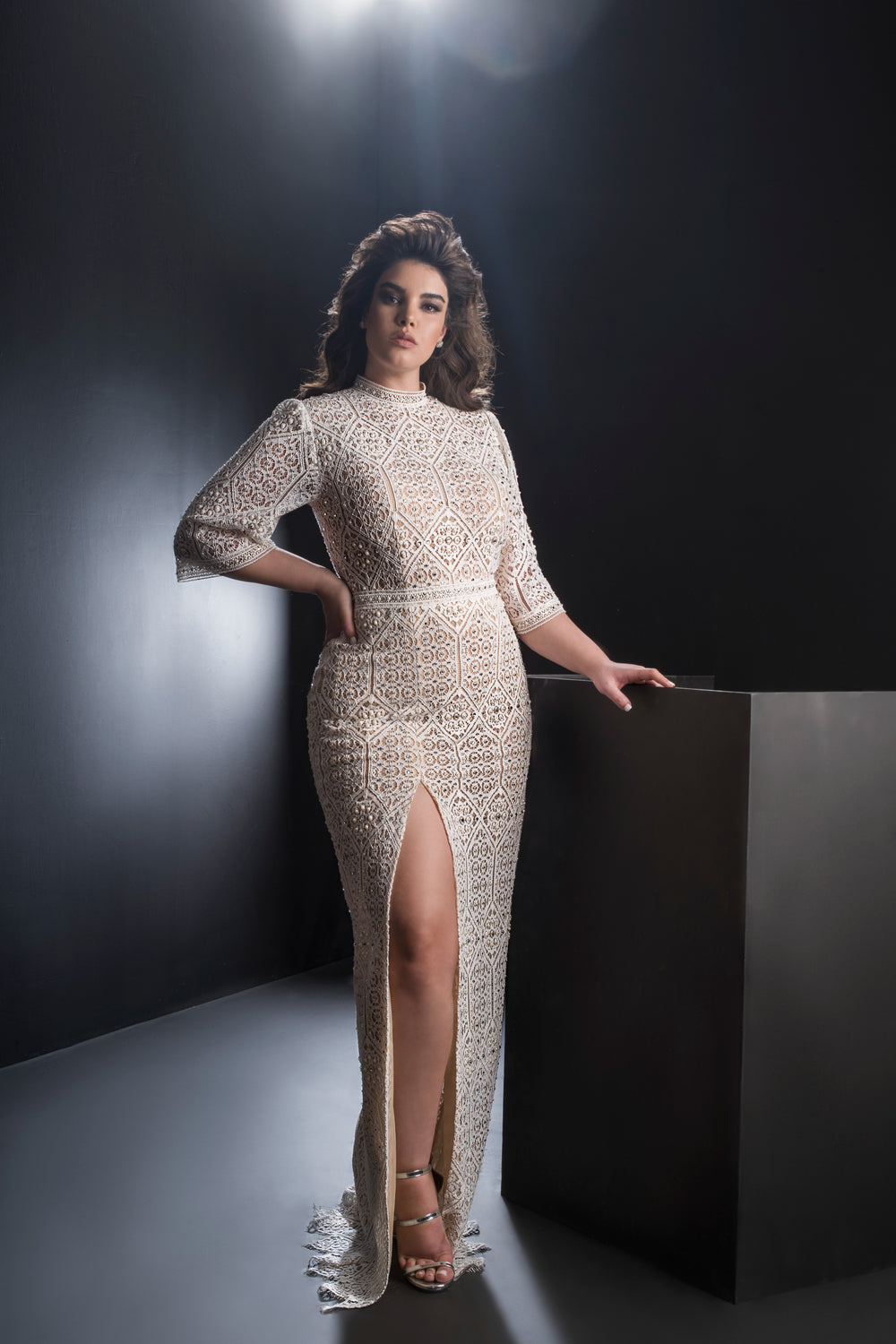 אישה עם שיער גלי מדגמנת את קולקציית Bridal Plus 2019 - שמלת Vienna, שמלה ארוכה עם דוגמת תחרה וחתך גבוה. היא פוזלת בביטחון, יד אחת על מותנה והשנייה על כן שחור בסטודיו מינימליסטי.