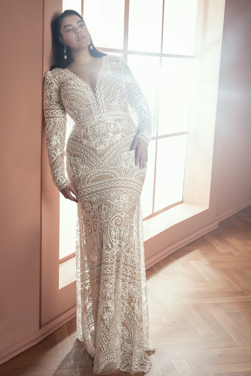 אישה בשמלת Kiana מקולקציית Bridal Plus 2021 עומדת ליד חלון מואר בשמש, נשענת על קיר בצבע אפרסק על רצפת עץ, ומדגימה את האלגנטיות של שמלות כלה.