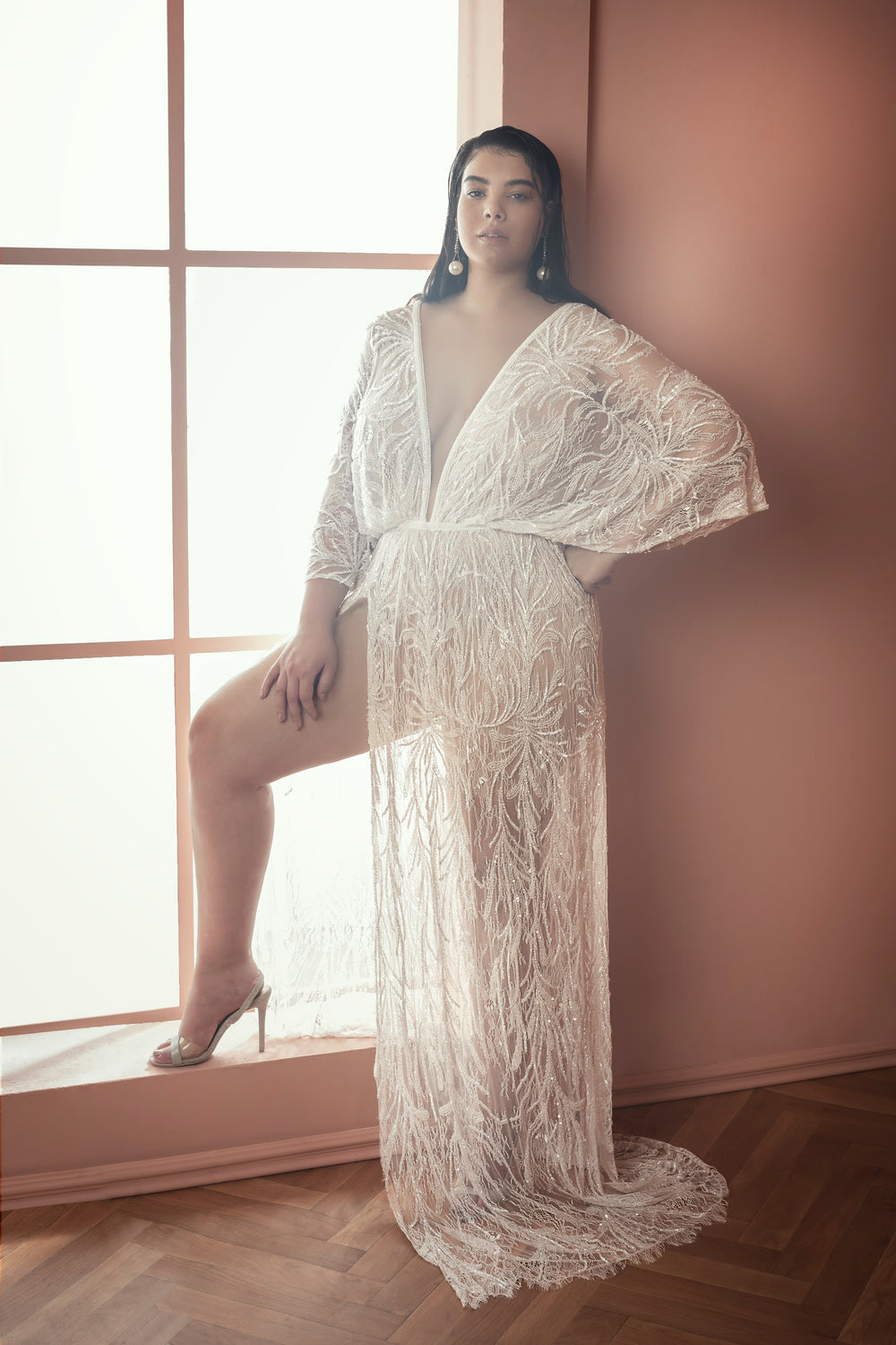 אישה לבושה בשמלה ארוכה, לבנה, שקופה ומרוקמת מקולקציית Bridal Plus 2021 - Mikaela עומדת בביטחון ליד חלון גדול בחדר מואר באור רך, בגווני אפרסק, עם רצפות עץ.