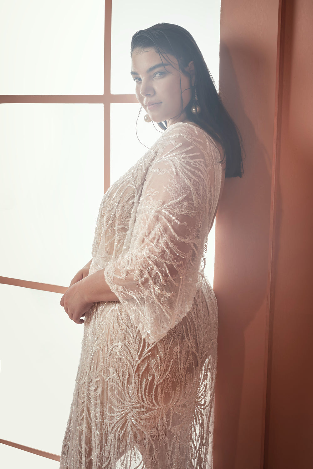אישה עם שיער ארוך וכהה עומדת ליד חלון בשמלה אלגנטית וטהורה בצבע לבן "Bridal Plus Collection 2021 - Mikaela". מוארת באור טבעי, היא מביטה בעדינות למצלמה, פרופיל הצד שלה שלו ואלגנטי.