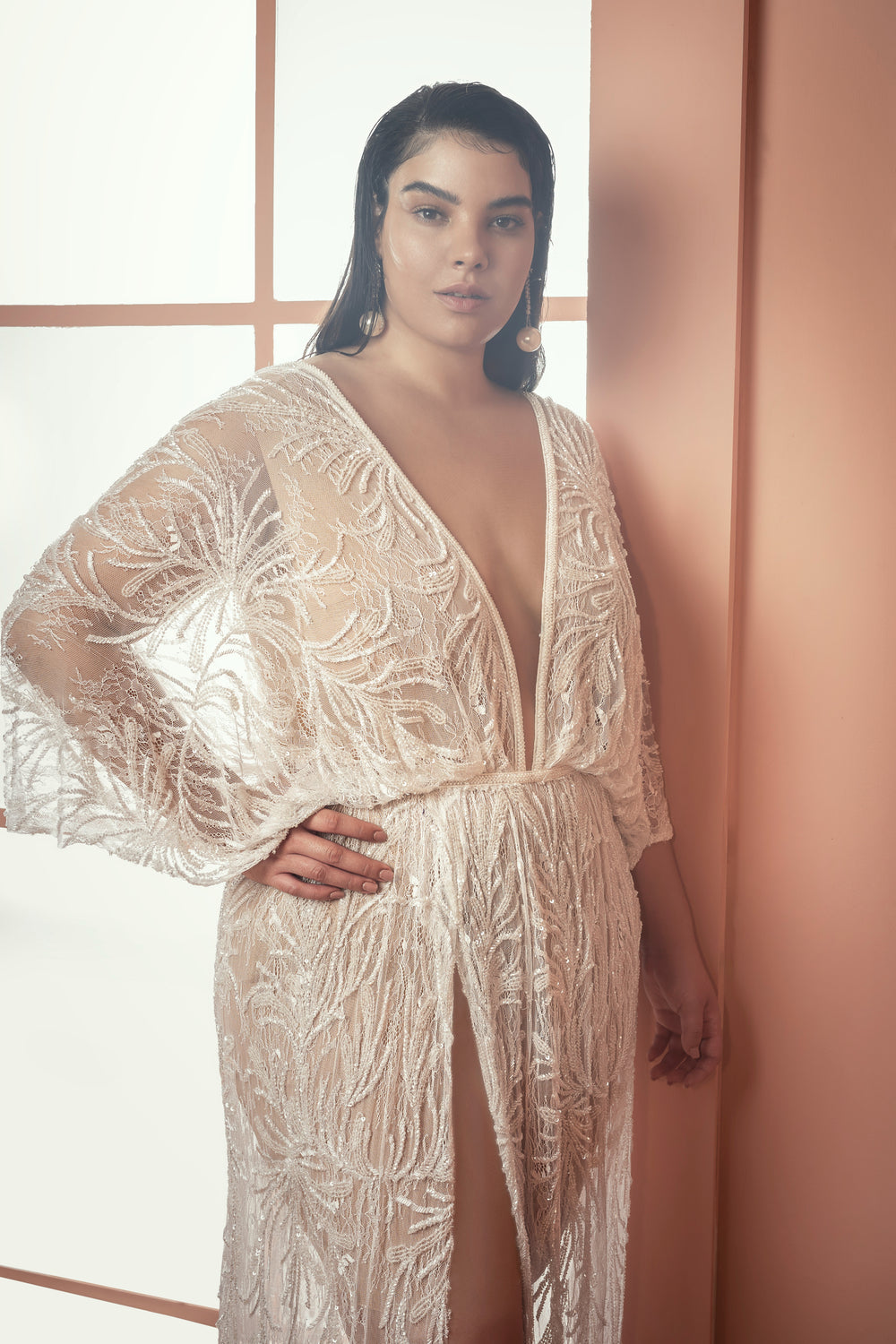 אישה עם שיער ארוך וכהה פוזלת בביטחון בשמלת Mikaela הלבנה והשקופה מקולקציית Bridal Plus 2021, עם רקמה מורכבת, מחשוף V עמוק ושרוולים רחבים על רקע קיר אפרסק וחלונות חלבית.