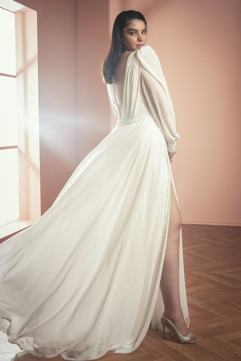 אישה מקולקציית Bridal Plus 2021 - שמלת Mercy, עם שרוולים ארוכים, גב עמוק וחצאית שיפון, עומדת בתוך הבית על רצפת עץ. אור השמש מדגיש את נעלי העקב שלה ואת הבד הלבן האלגנטי כשהיא מביטה מעבר לכתפה.