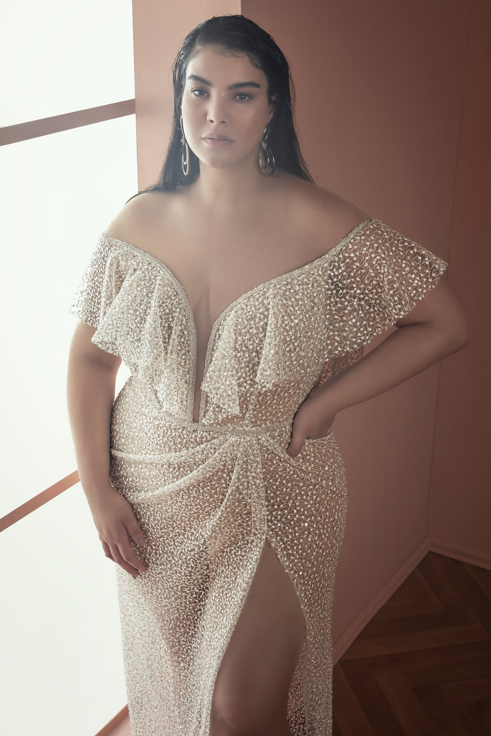 אישה עם שיער ארוך וכהה לובשת את שמלת הכלה הזוהרת Bridal Plus Collection 2021 - Aya, בעיצוב שקוף עם כתפיים חשופות, מחורר בחרוזים, עם מחשוף עמוק וחתך גבוה, עומדת בחדר מואר באור רך עם קירות בצבע אפרסק ורצפת עץ.