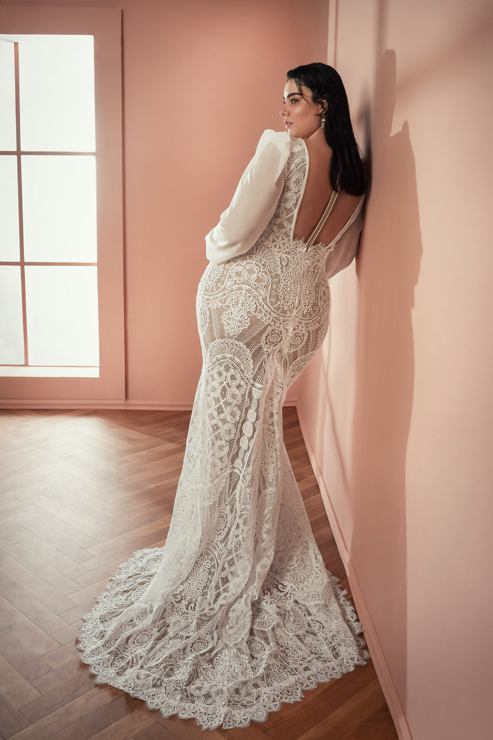 אישה לובשת שמלת כלה מהקולקציה Bridal Plus Collection 2021 - שמלת כלה מתחרה עם שרוולים ארוכים וגב פתוח, עומדת על רצפת עץ מול קיר ורוד, מביטה מעבר לכתפה בזמן שאור השמש חודר מבעד לחלון גדול.