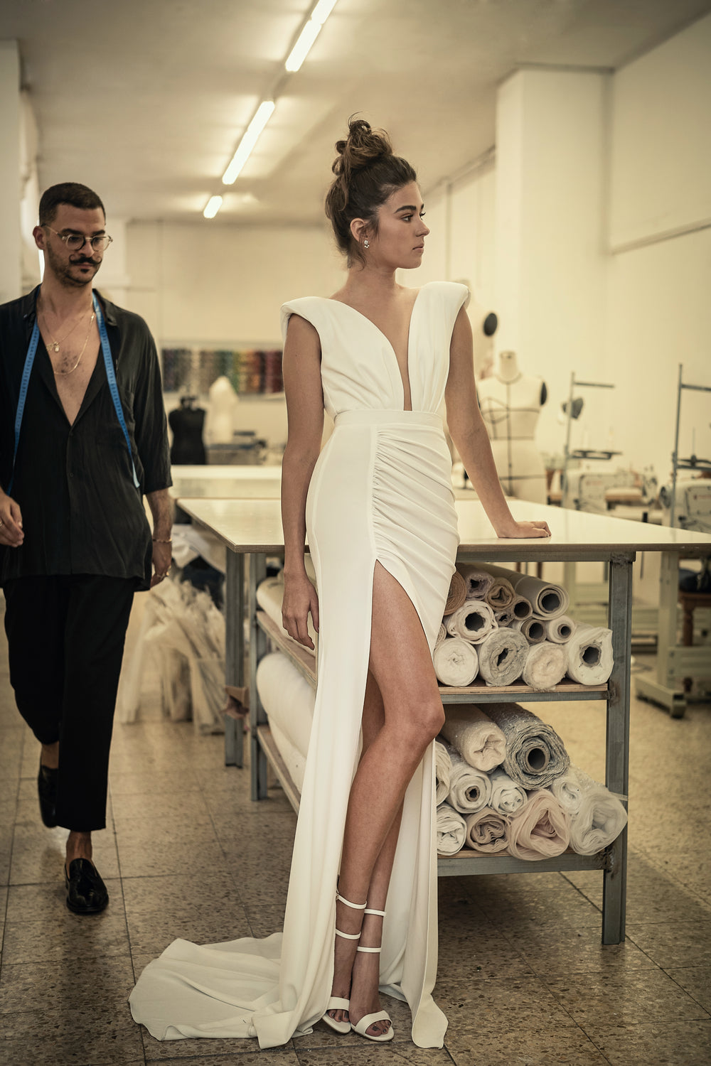 אישה לבושה בשמלת כלה מחויטת מקולקציית הכלות 2021 - Lilly שמלת כלה עם חתך גבוה נשענת על שולחן עבודה מכוסה בבד בסטודיו לתפירה, בעוד גבר עם משקפיים ומטר מדידה הולך ברקע.