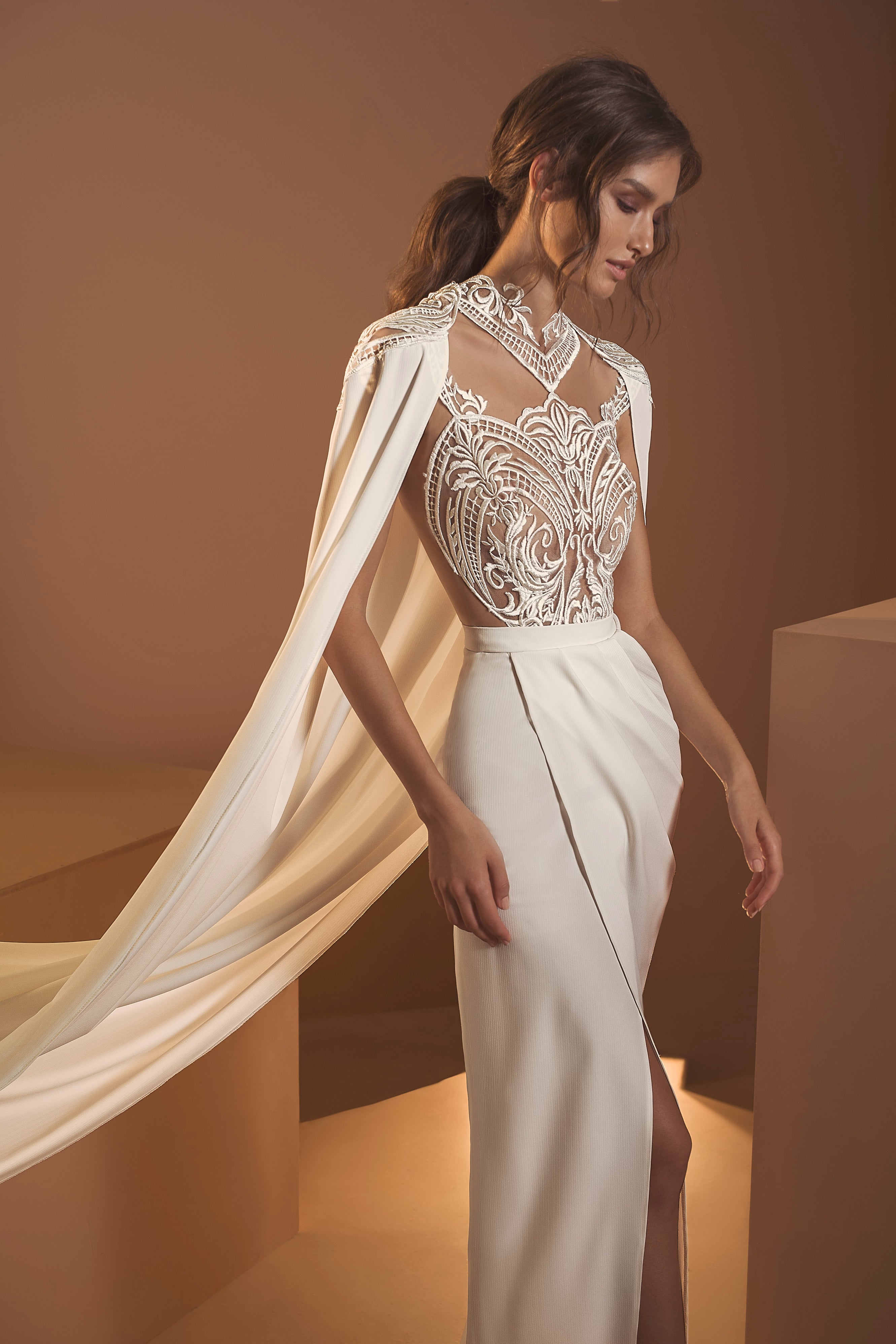 אישה בשמלת "אריאל" האלגנטית מקולקציית הכלות 2020 של דרור קונטנטו, עם תחרה מורכבת ושרוולים זורמים דמויי גלימה, עומדת על רקע מואר באור רך, שיערה גלי וזורם – ומגלמת את מהות שמלות כלה.