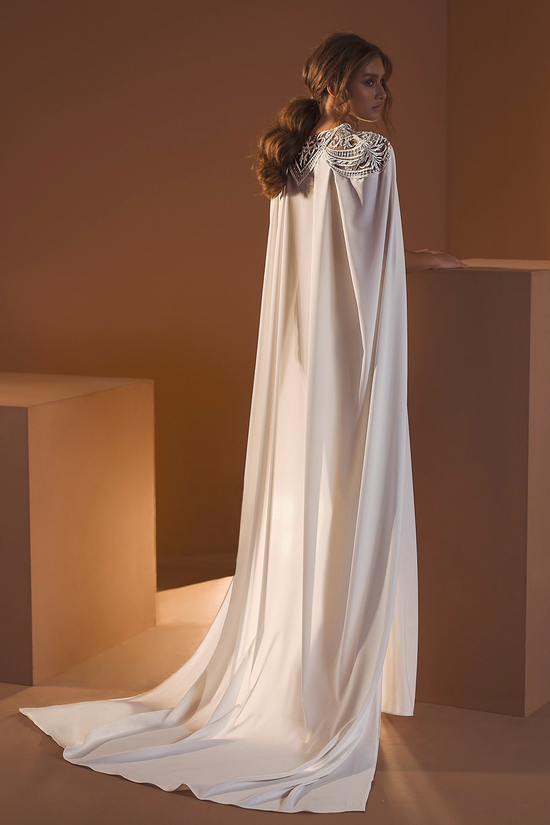 אישה עם שיער חום גלי עומדת בחדר מינימליסטי לבושה בשמלת "אריאל" מקולקציית הכלות 2020 של דרור קונטנטו, עם כתפיים מעוטרות ושובל דמוי גלימה. היא פונה עם גבה, מניחה את ידה על בלוק בצבע בז'.