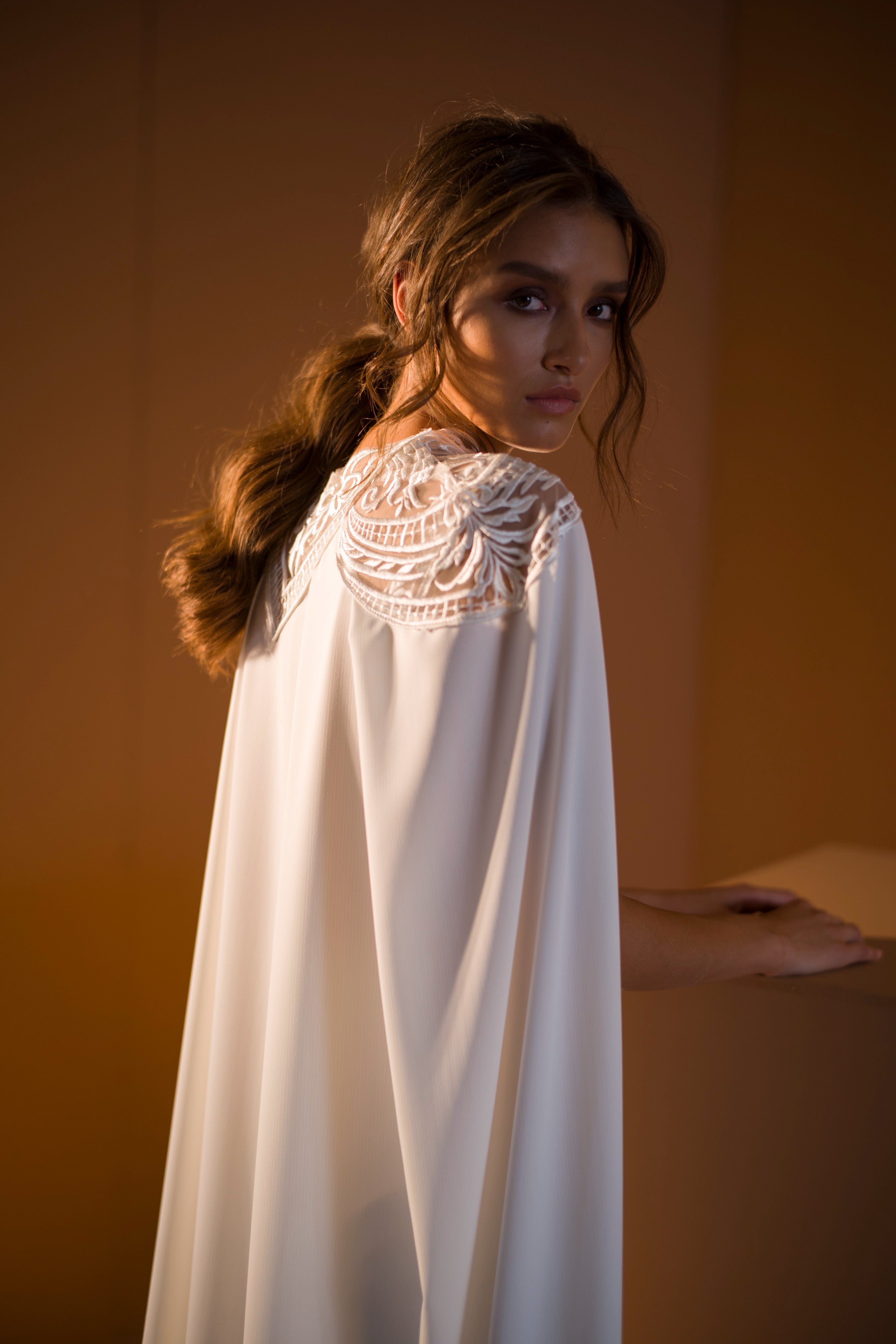 אישה עם שיער חום גלי, בשמלת אריאל מקולקציית הכלות 2020, עם תחרה מורכבת בגב, עומדת באור רך וחם ומביטה מעבר לכתפה.