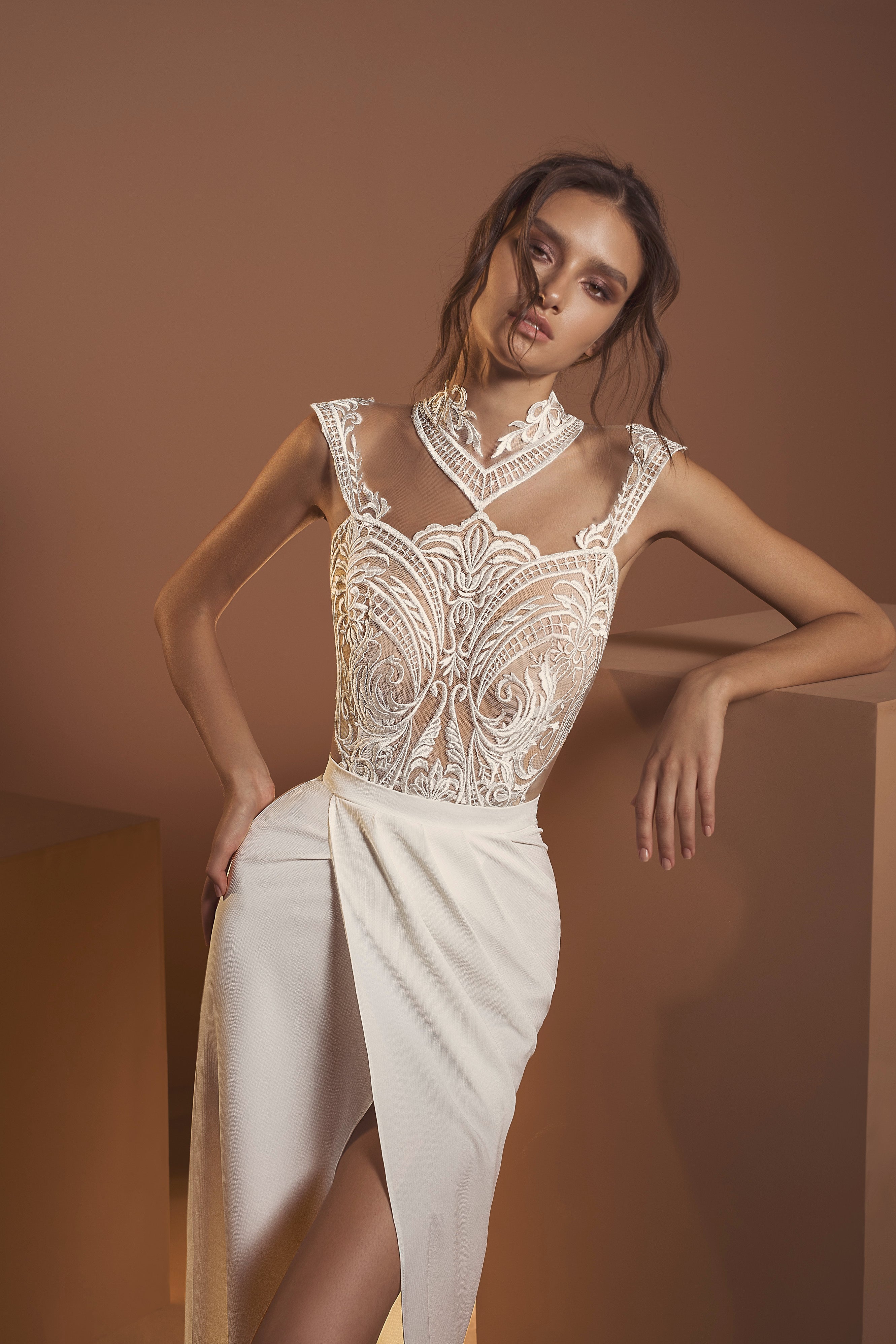 אישה בשמלת "אריאל" הלבנה והאלגנטית מקולקציית הכלות 2020 של דרור קונטנטו, עם מחוך תחרה מורכב ומחשוף שקוף, מצטלמת על רקע חום ניטרלי, נשענת על כן.