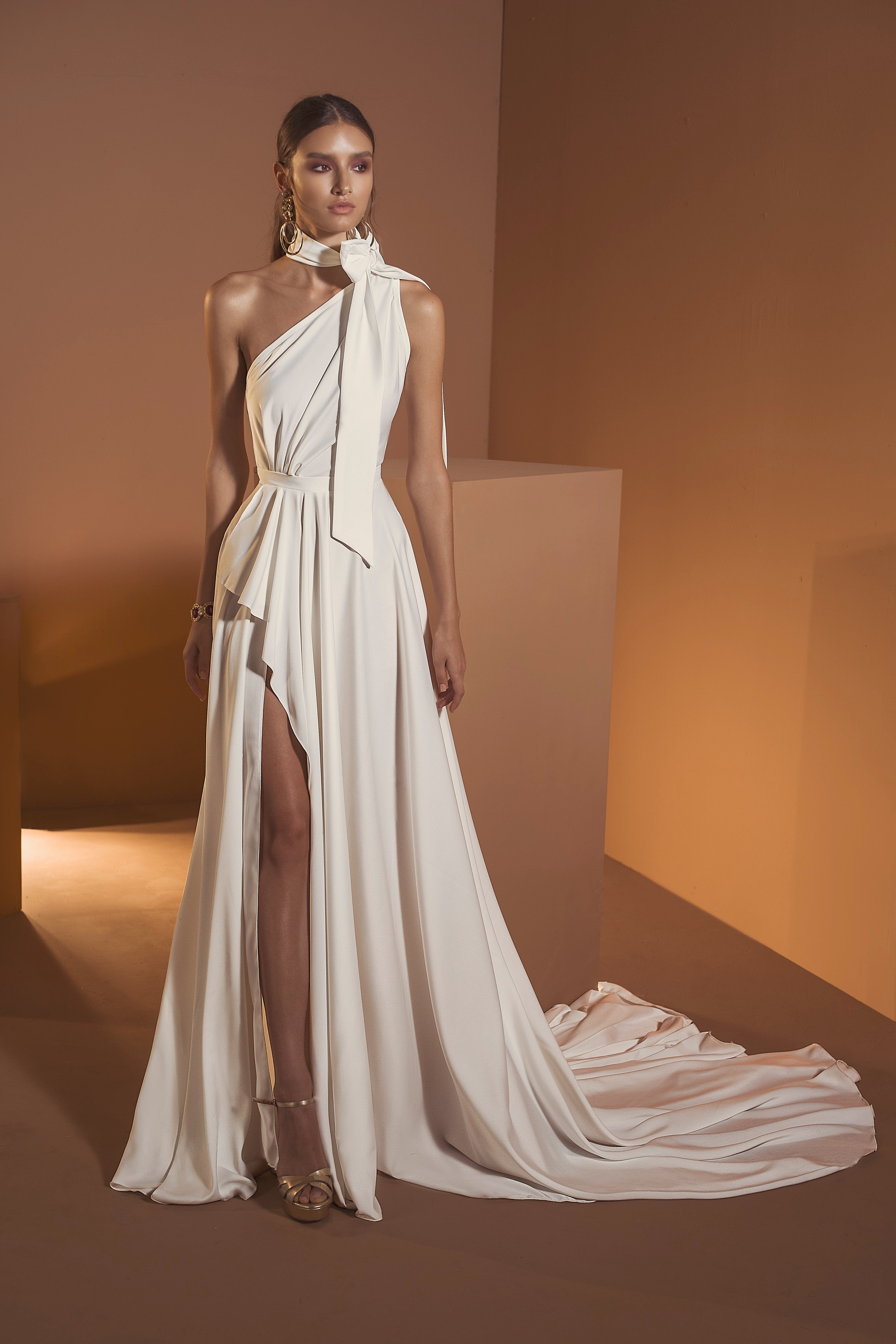 בחדר בצבע שמנת, אישה לובשת את שמלת אריאל מקולקציית הכלות 2020 של דרור קונטנטו — שמלה לבנה וזורמת עם כתף אחת, חתך גבוה, שובל ארוך, קשת בצוואר, עגילי זהב ונעלי עקב עם רצועות.