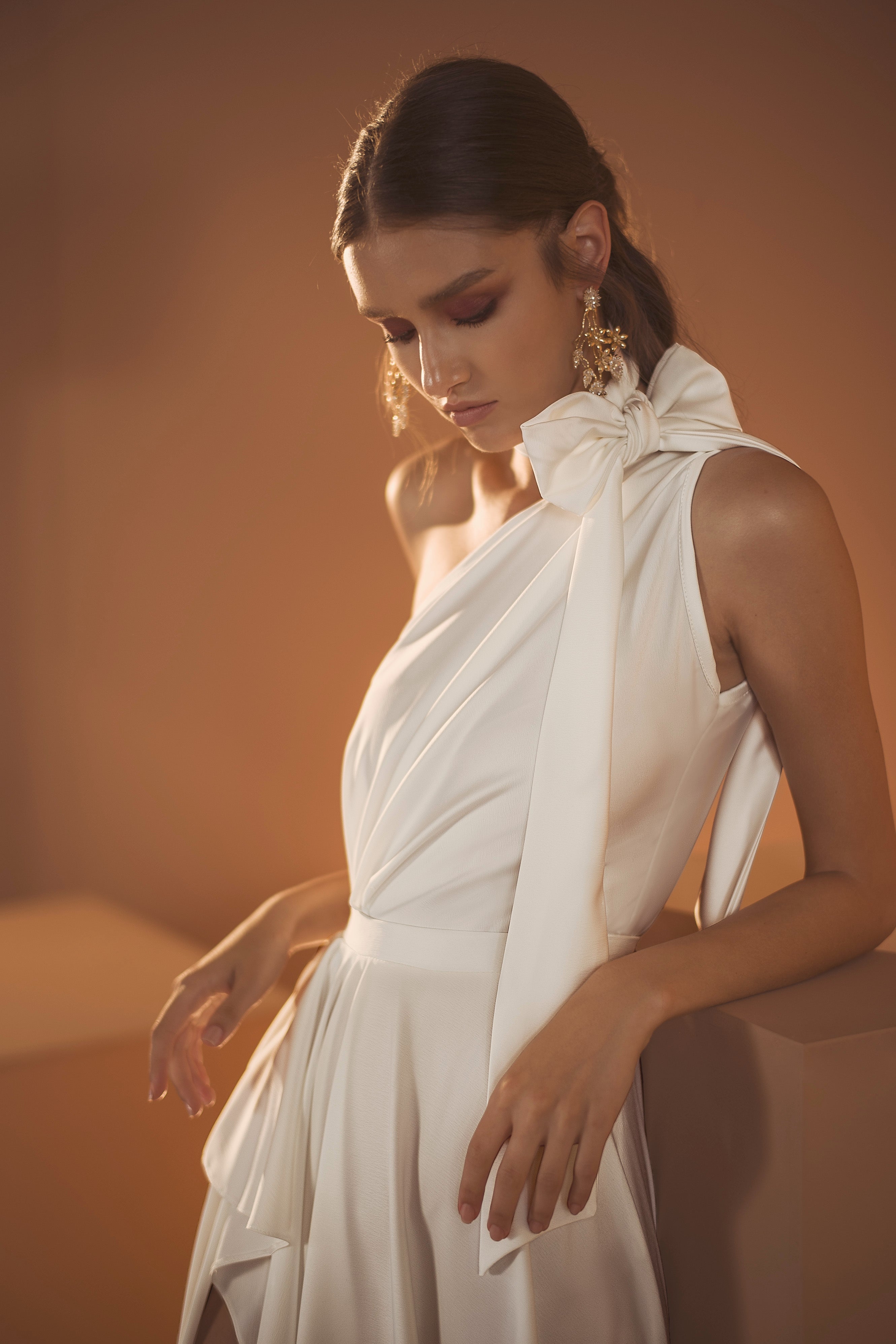 אישה בשמלת אריאל מקולקציית הכלות של דרור קונטנטו לשנת 2020, בעלת עיצוב אלגנטי עם כתף אחת, מצטלמת עם שיער חום גלי ועגילי סטטמנט על רקע חם ומואר באור רך.