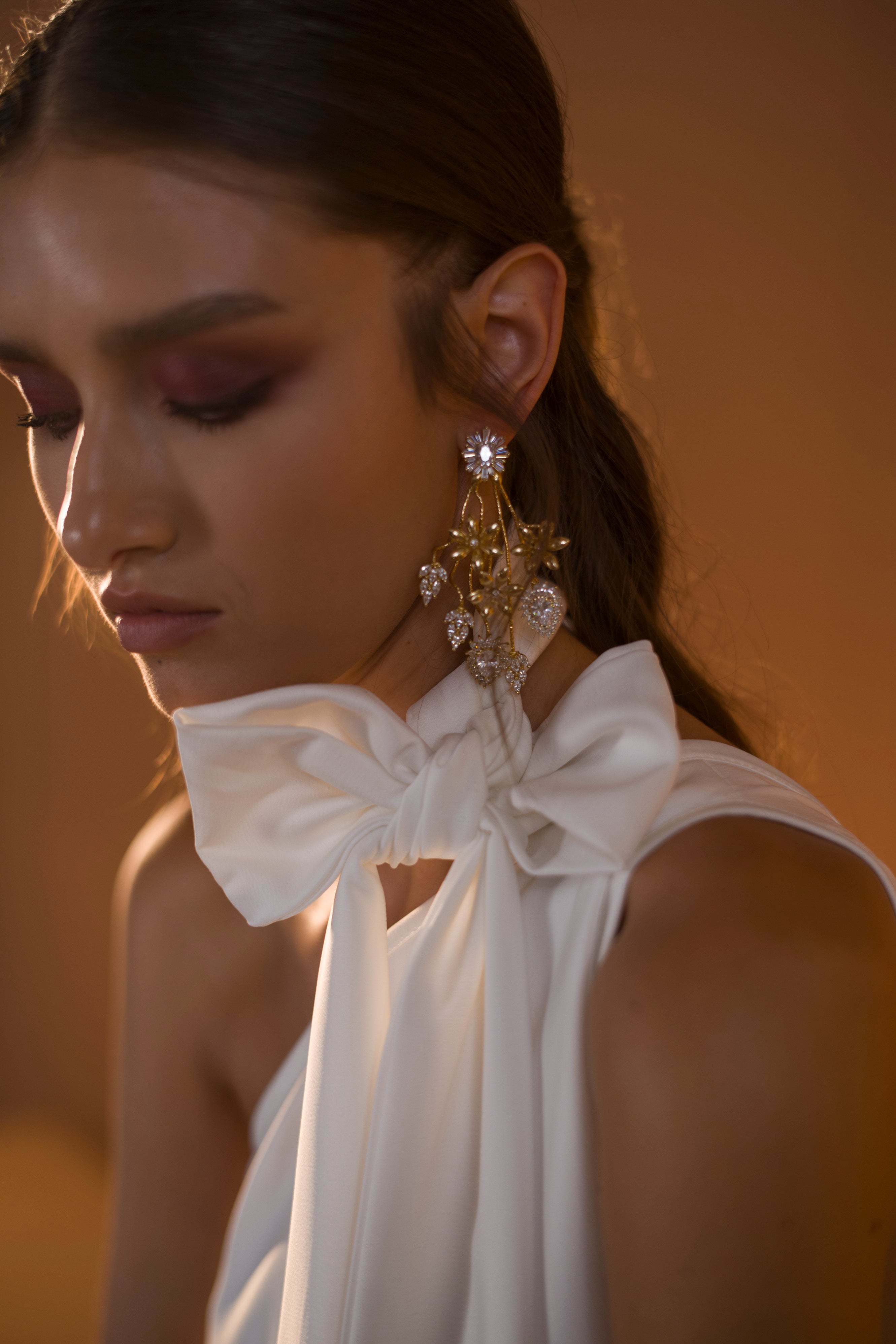 אישה לבושה בשמלה לבנה ללא שרוולים מקולקציית הכלות 2020 - אריאל של דרור קונטנטו, עם קשת גדולה על הכתף, עונדת עגילי זהב ויהלומים מעוטרים. שיערה החום מסורק לאחור, עם איפור עיניים סגול דרמטי על רקע מטושטש ורך.