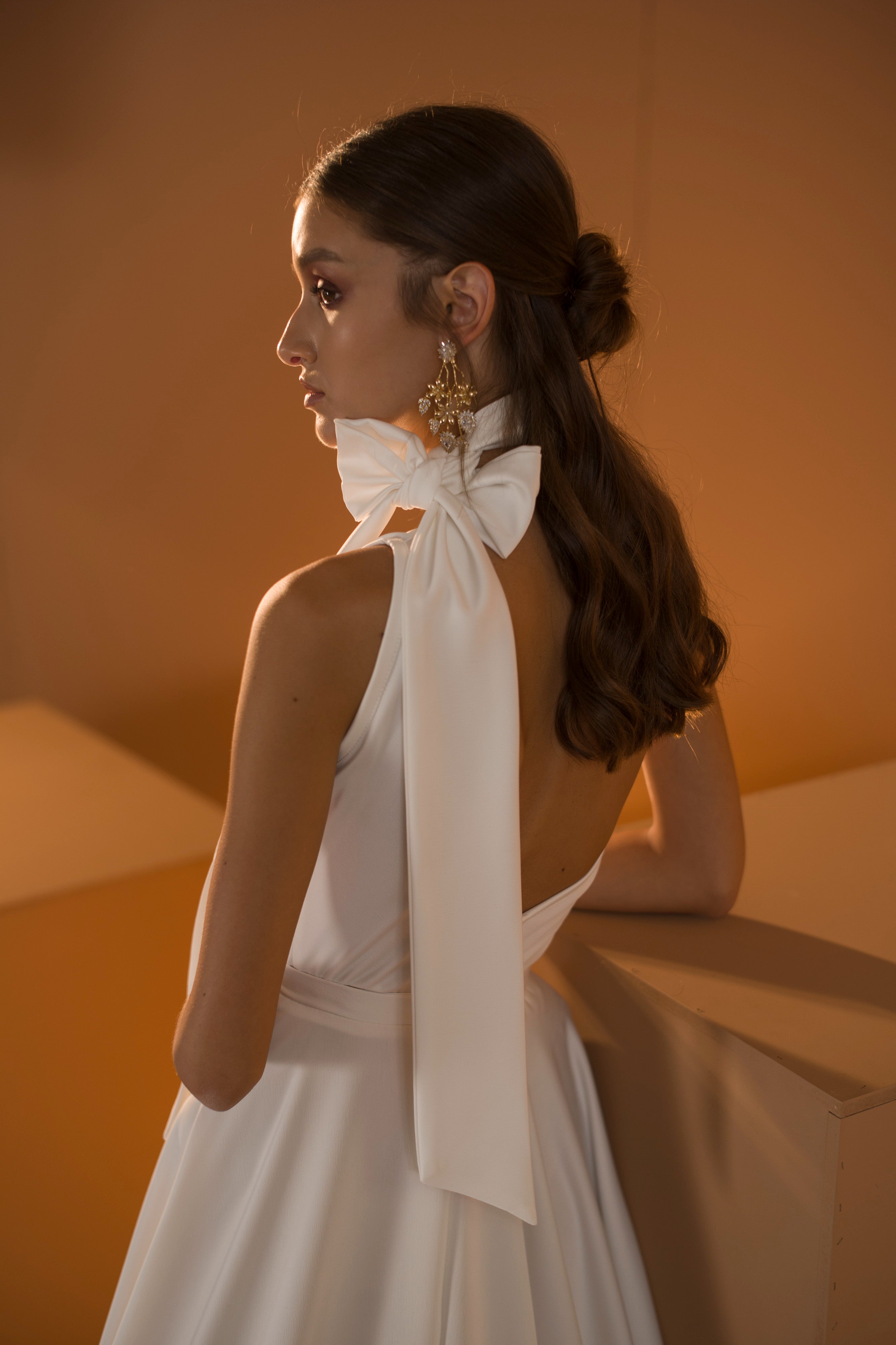 אישה לבושה בשמלת כלה מקולקציית 2020 - אריאל, שמלת דרור קונטנטו לבנה עם קשת גדולה על הכתפיים, עומדת בחדר מואר באור חם. שיערה החום הארוך והגלי אסוף בחלקו, והיא עונדת עגילים מתנדנדים ומקושטים.