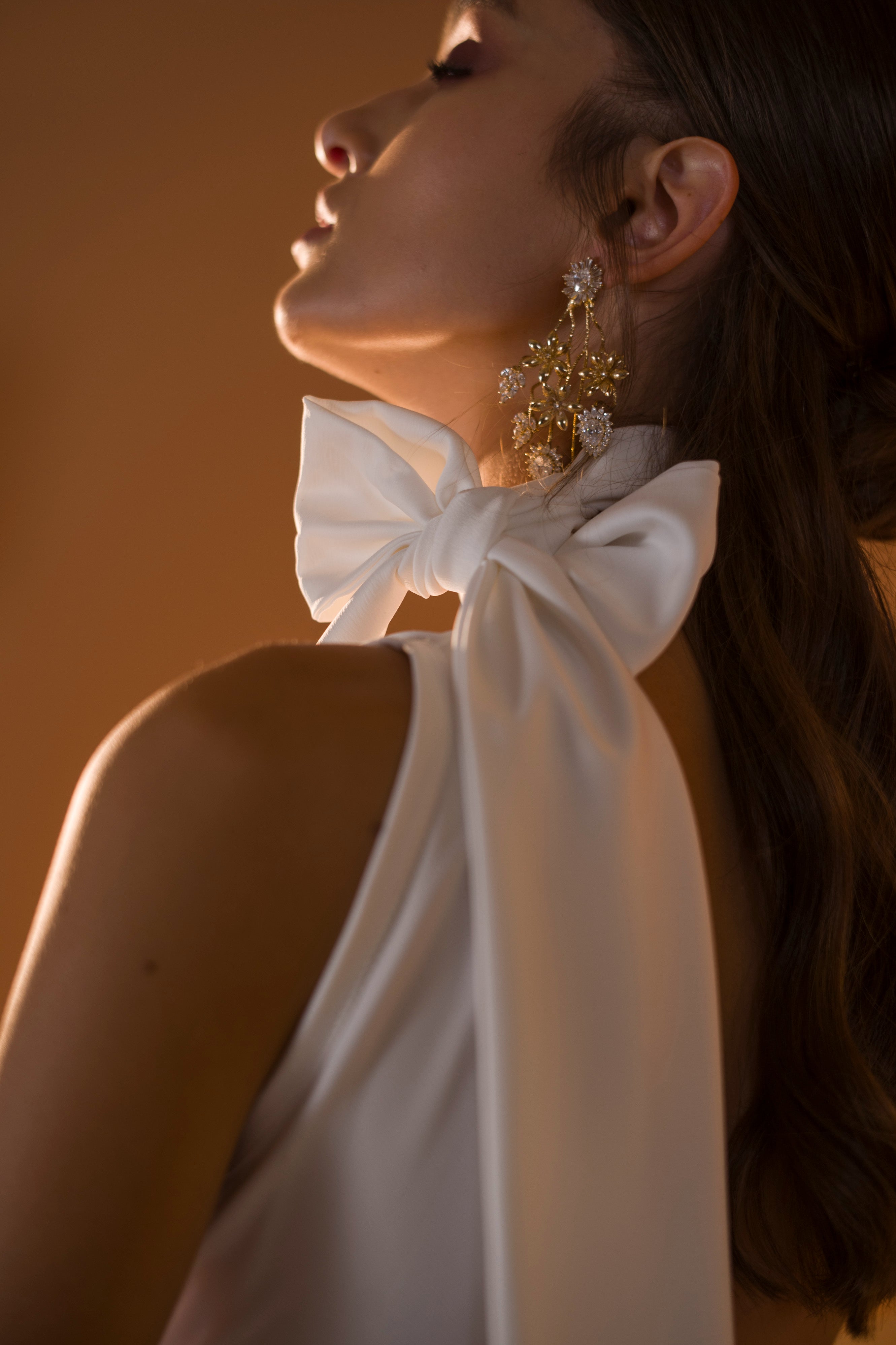 אישה בפרופיל לובשת שמלה לבנה עם קשת גדולה על הכתף ועגילי זהב מעוטרים מקולקציית הכלות 2020 של דרור קונטנטו - אריאל, שיערה החלק ותנוחתה האלגנטית מודגשים על ידי תאורה רכה וחמימה.