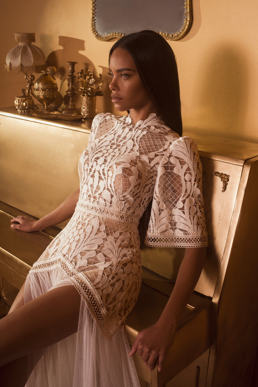 אישה לבושה בשמלת בלייר מקולקציית הכלות 2019 יושבת על פסנתר זהוב בחדר מואר באור חם, המעוטר באלמנטים וינטג'יים מברזל ומראה קיר.