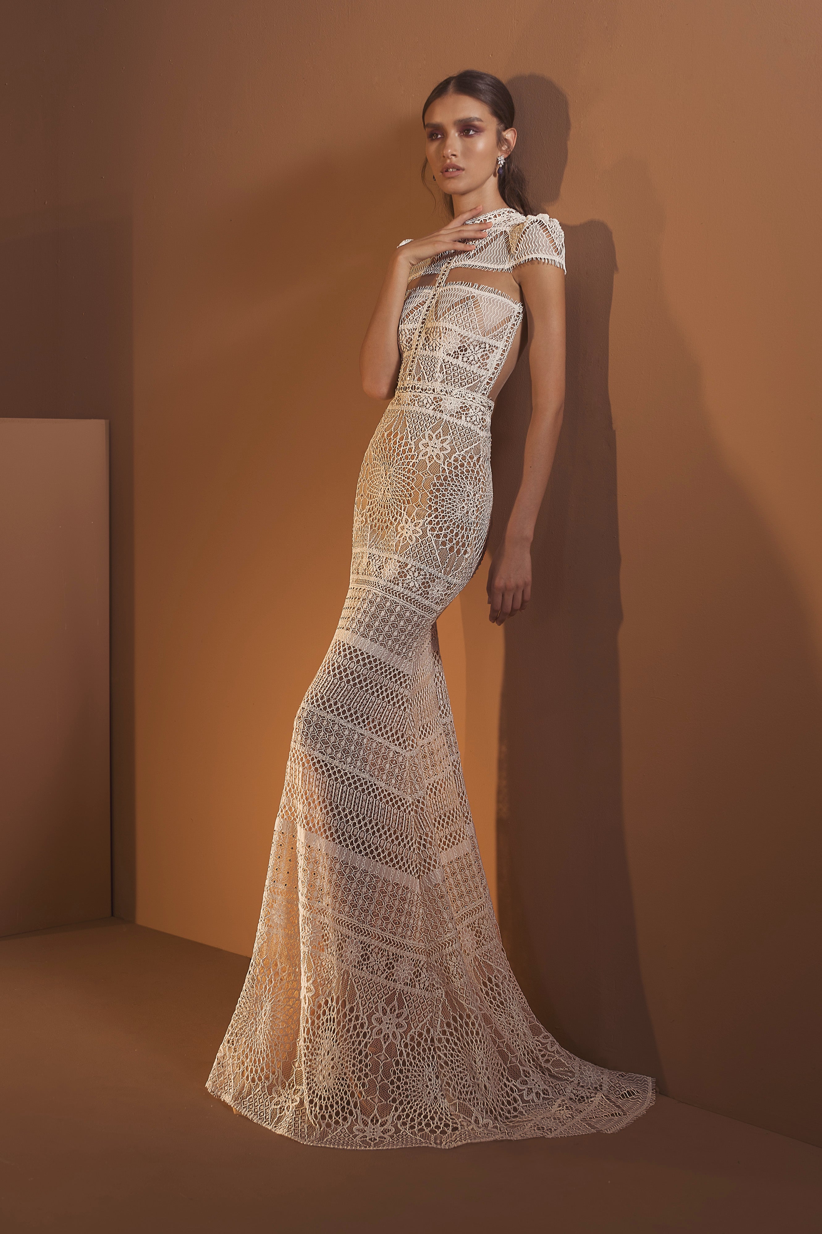אישה מצטלמת על רקע קיר חום בשמלת דניאלה מקולקציית הכלות 2020 של דרור קונטנטו, עם גזרה צמודה באורך מלא, שרוולים קצרים, דוגמאות תחרה, פרטים מורכבים בסריגה וחתך מתרחב למראה אלגנטי.