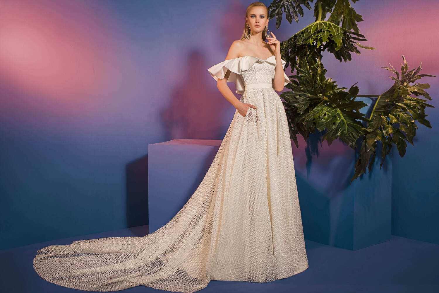 אישה לבושה בשמלת כלה מקולקציית הכלות - Pure - 04, שמלת טקסטורה לבנה עם כתפיים חשופות, עומדת על רקע סגול וכחול עם עלים ירוקים גדולים, מביטה קדימה וידה האחת קרובה לפניה.