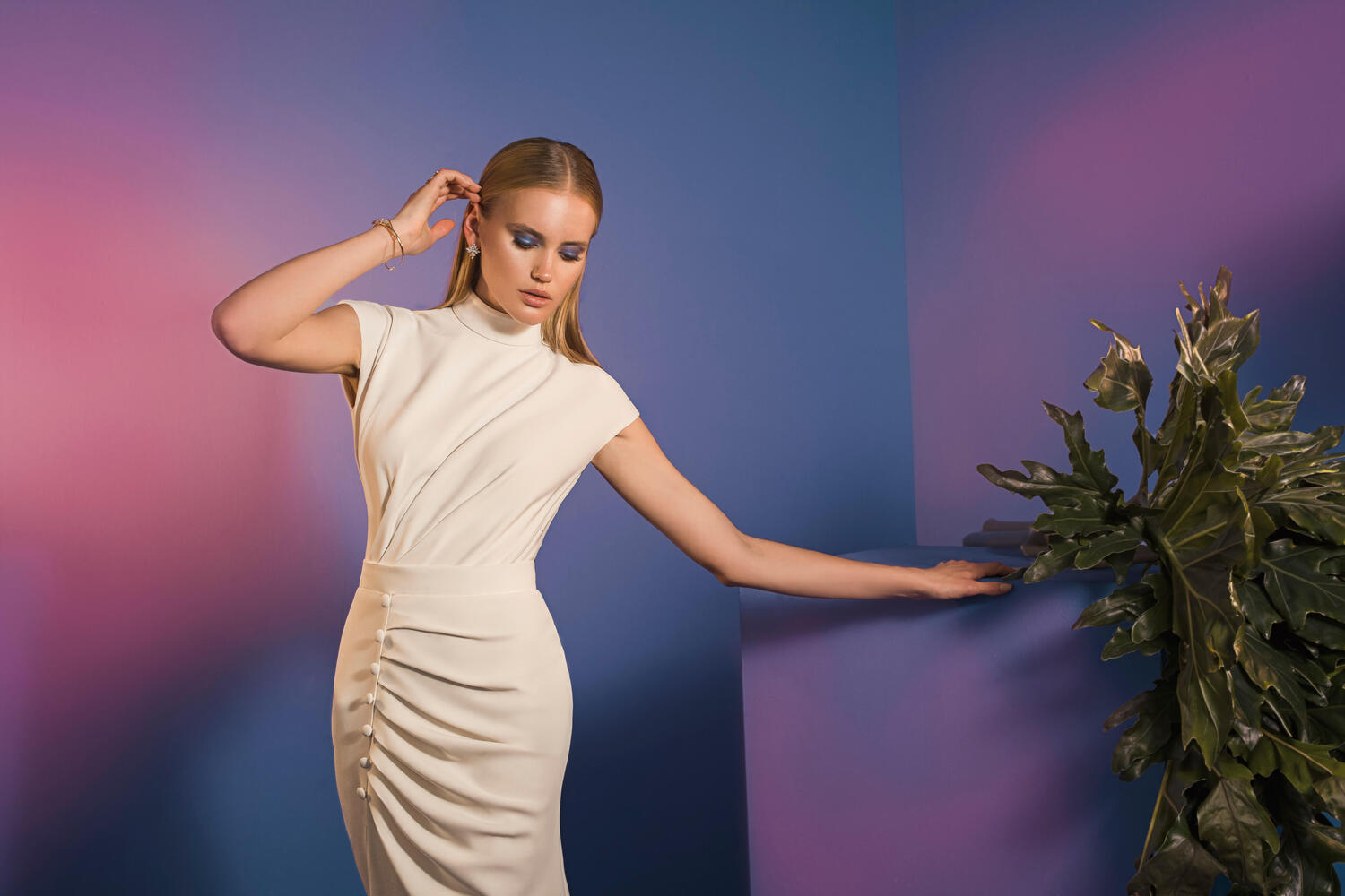 אישה בשמלת כלה לבנה מקולקציית הכלות - Pure - 08 מצטלמת ליד צמח עלים, יד אחת נוגעת בשיערה, והשנייה על כן, על רקע כחול וסגול עם תאורה ורודה וסגולה רכה.