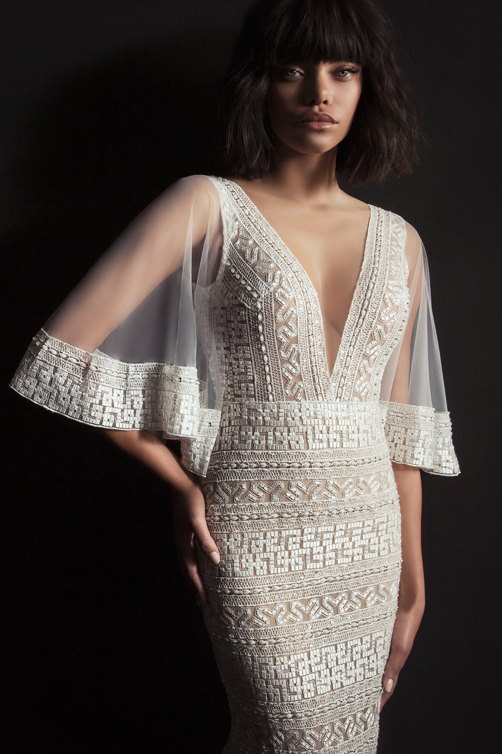 אישה עם שיער קצר, גלי וכהה מדגמנת את שמלת Karina מקולקציית הכלות 2017, המציגה רקמה גיאומטרית מורכבת ושרוולים שקופים ומתרחבים על רקע כהה.
