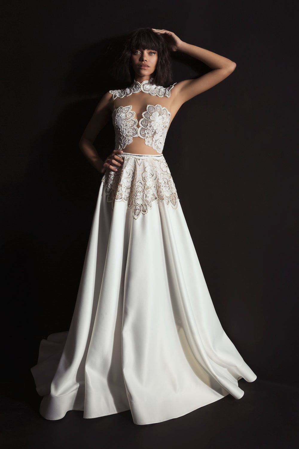 אישה עומדת על רקע שחור בשמלת Kori מקולקציית הכלות 2017 של דרור קונטנטו, שמלה לבנה באורך הרצפה עם אפליקציות תחרה על המחשוף והכתפיים — בחירה אלגנטית לשמלות כלה.