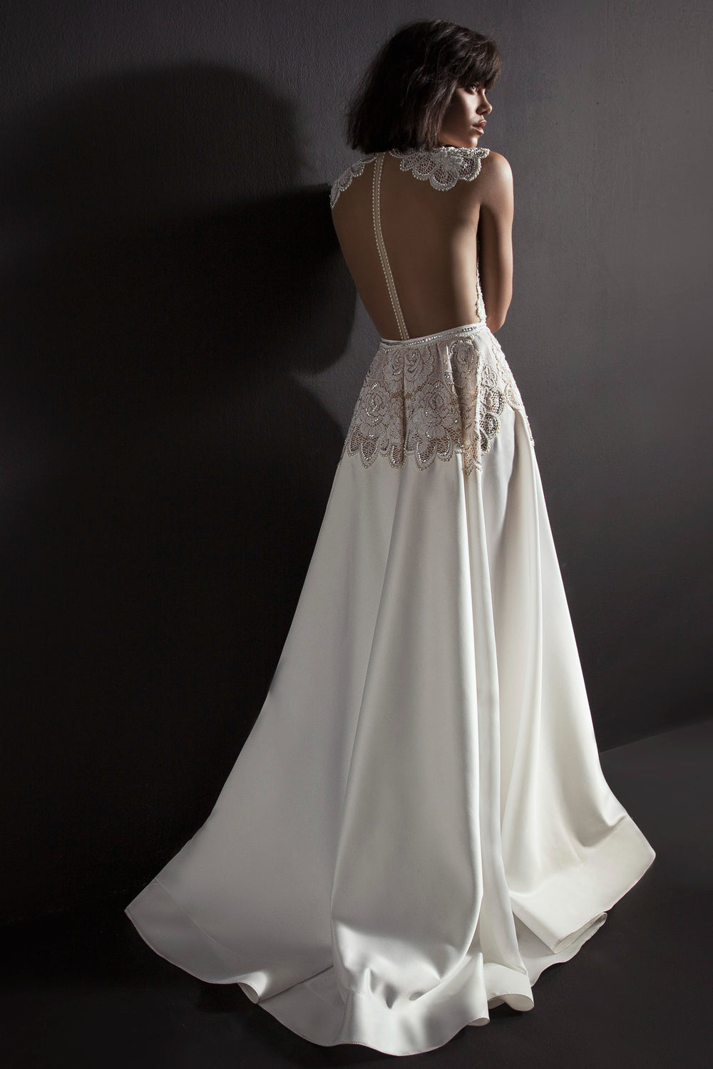 אישה בשמלת Kori מקולקציית הכלות 2017 של דרור קונטנטו מצטלמת על רקע כהה, ומציגה את פרטי התחרה, החצאית הזורמת והרצועות המעוטרות באבנים יקרות בגב הפתוח.