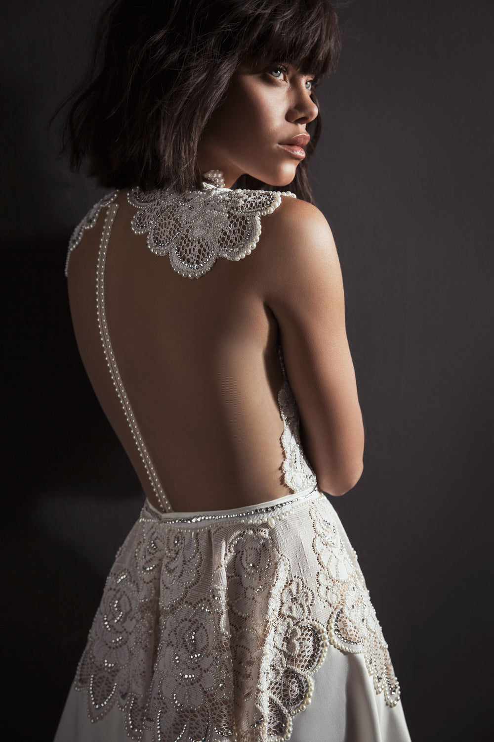 אישה עם שיער קצר וכהה לובשת את שמלת Kori מקולקציית הכלות 2017, עם תחרה מורכבת ופרטי פנינה, על רקע כהה, תוך שהיא מביטה לצד.