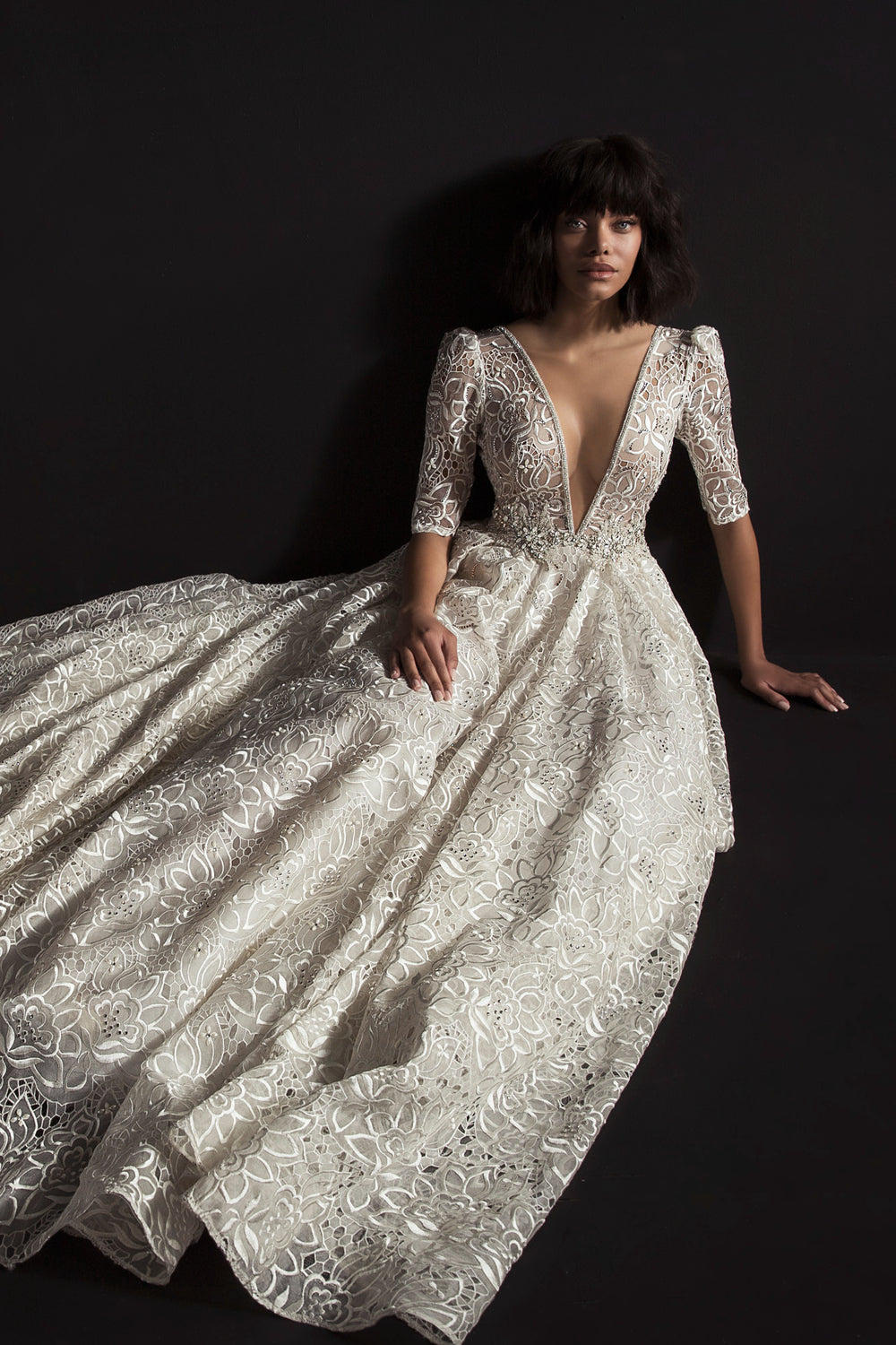 אישה עם שיער קצר וכהה יושבת על רקע שחור, לבושה בשמלת איירין מקולקציית הכלות 2017 של דרור קונטנטו — שמלת תחרה לבנה וזורמת עם מחשוף V עמוק ושרוולים אלגנטיים באורך המרפק.