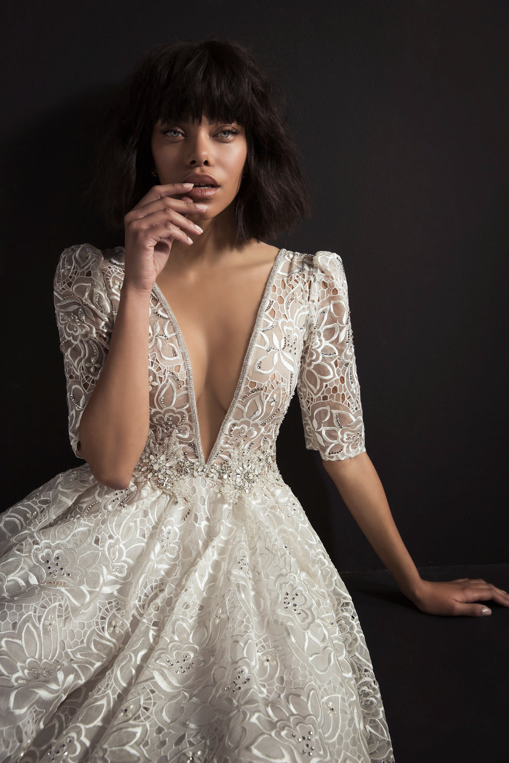 אישה לבושה בשמלת איירין מקולקציית הכלות 2017, עם תחרה, מחשוף V עמוק ושרוולים שקופים באורך חצי, יושבת על רקע כהה, מביטה קדימה וידה ליד פיה.