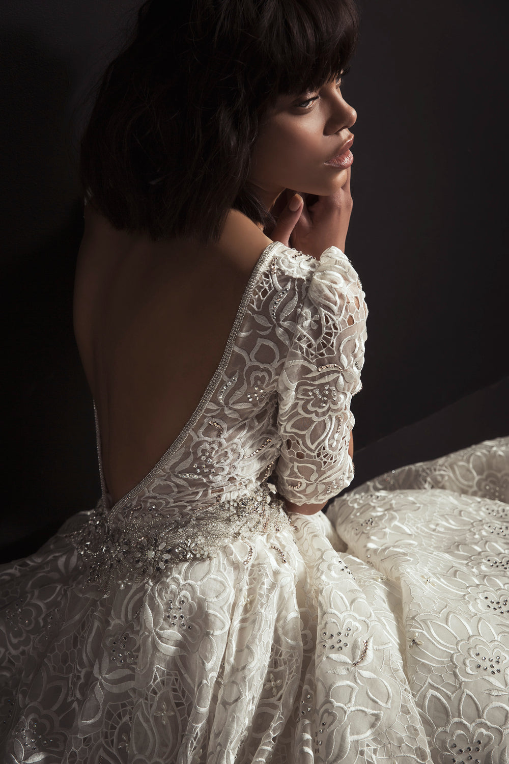 אישה עם שיער קצר וכהה, לבושה בשמלת "אירין" אלגנטית עם גב פתוח מתחרת, מקולקציית הכלות 2017, יושבת על רקע כהה ומביטה בצד במבט מהורהר.