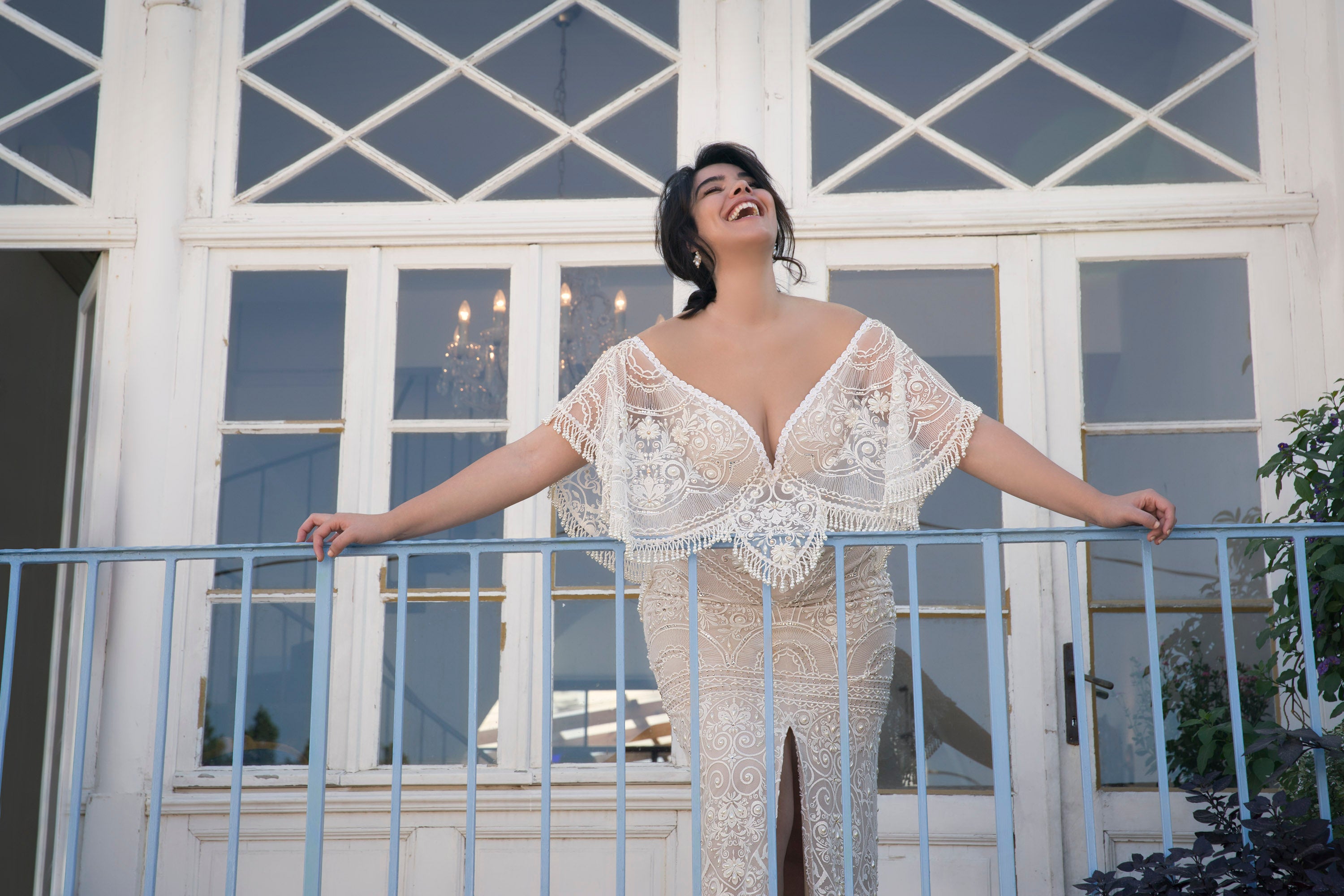 אישה בשמלת תחרה מנילה מקולקציית Bridal Plus 2017 עומדת במרפסת, זרועותיה פרושות, מחייכת ומביטה למעלה. מאחוריה, חלונות גדולים עם מסגרות לבנות מעוטרים בדוגמאות גיאומטריות.