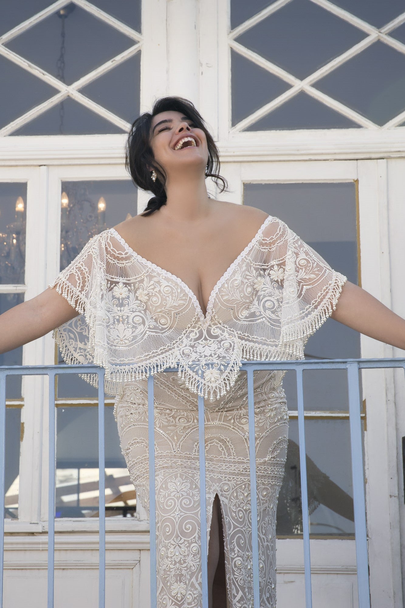 אישה בשמלה לבנה מתחרה עם כתפיים חשופות מקולקציית Bridal Plus 2017 - מנילה עומדת במרפסת, מחייכת וצוחקת עם הראש מוטה לאחור ליד דלתות זכוכית.