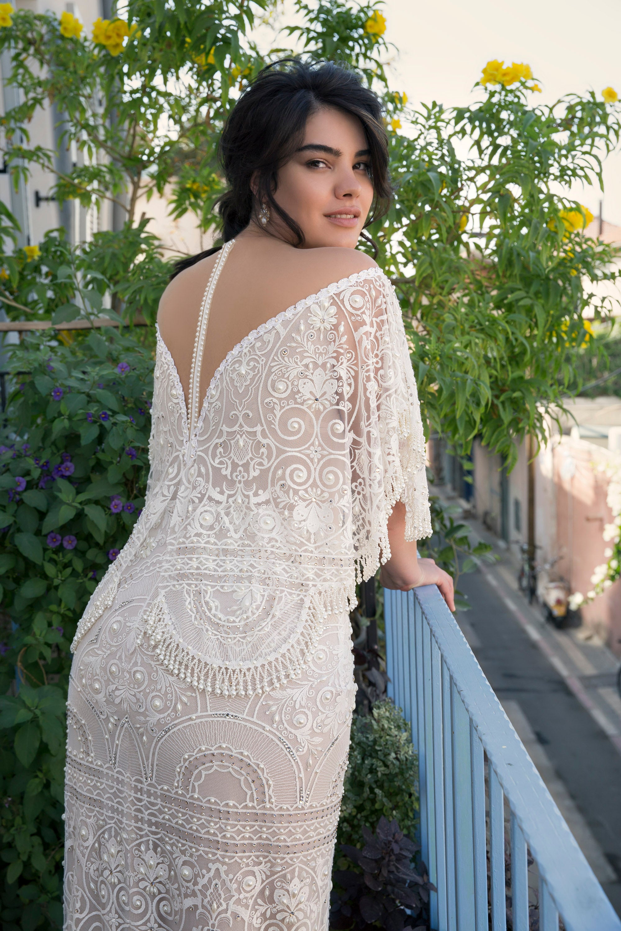 אישה לבושה בשמלה לבנה עם תחרה מקולקציית Bridal Plus 2017 - מנילה עומדת במרפסת, מחייכת מעבר לכתפה בין צמחים ירוקים ופרחים צהובים, עם רחוב עירוני ובניינים ברקע.