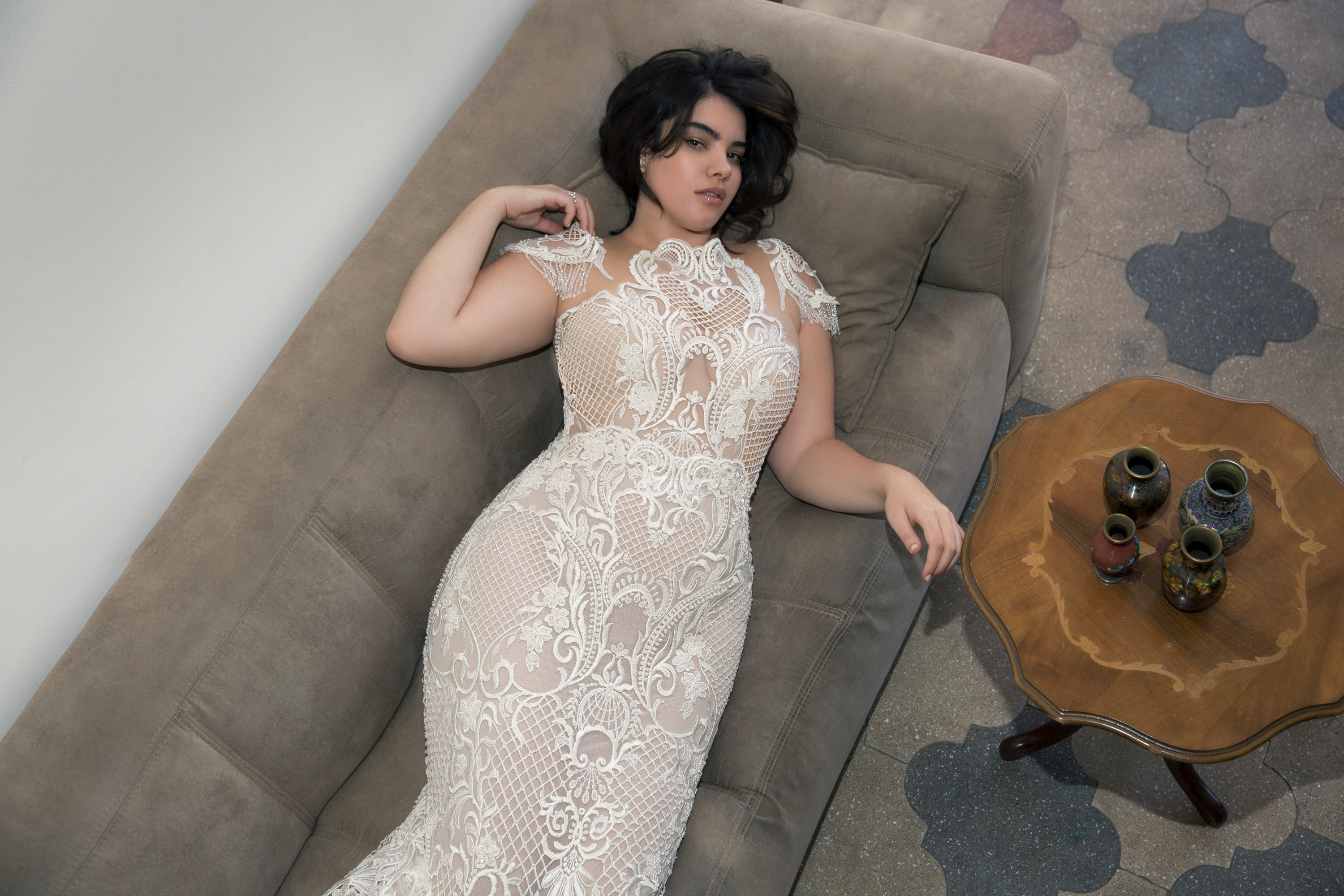 אישה לבושה בשמלת תחרה לבנה עם דוגמה מורכבת מקולקציית Bridal Plus 2017 - Adria מאת דרור קונטנטו שוכבת על ספה בצבע בז', מביטה למעלה, כשלידה שולחן עץ ואגרטלים דקורטיביים על רצפת אריחים מעוטרת.