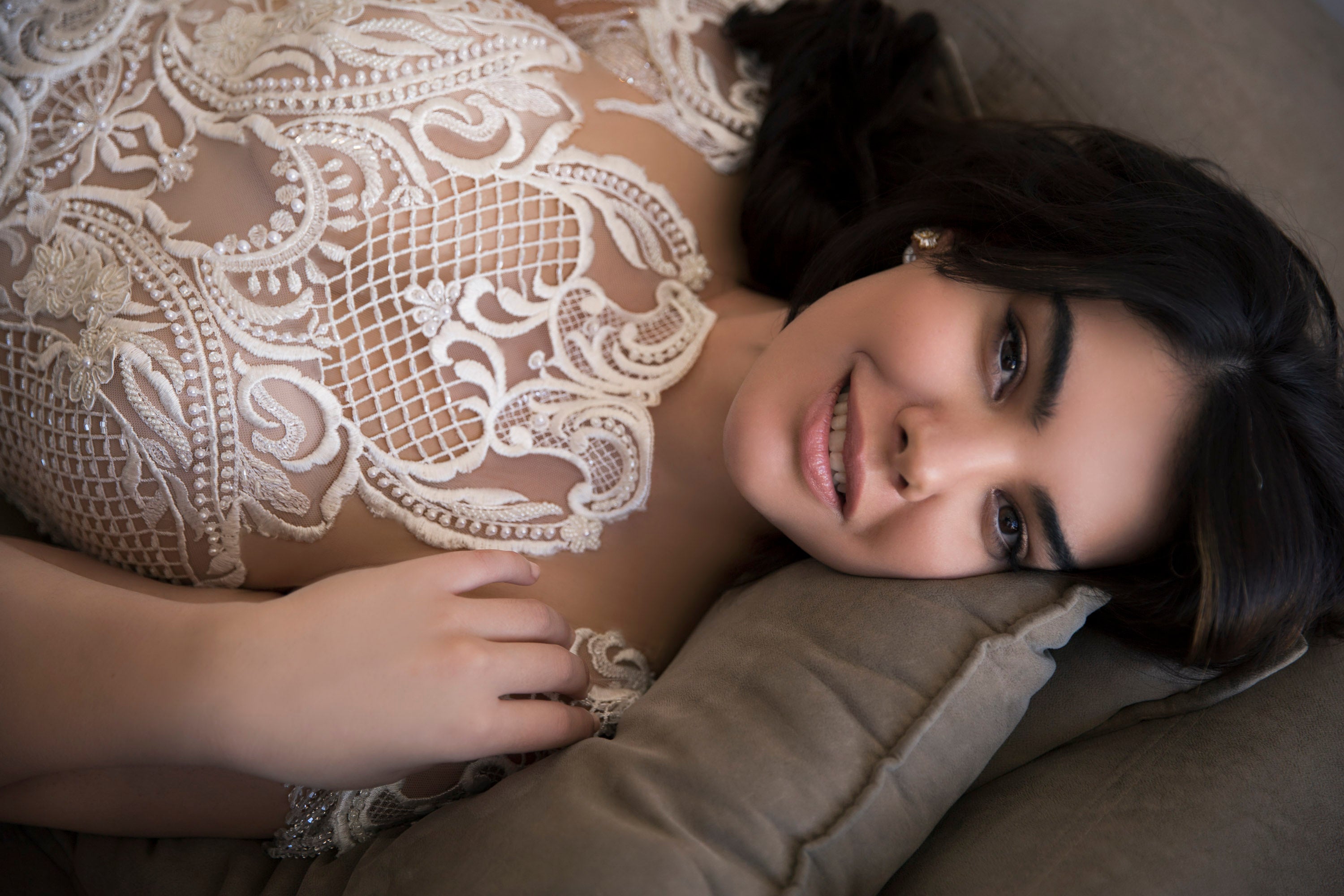 אישה עם שיער כהה שוכבת על כרית, מחייכת ברכות למצלמה כשהיא לובשת את שמלת Adria מקולקציית Bridal Plus 2017 של דרור קונטנטו. ידה מונחת ליד הצווארון, ומדגישה את דוגמת התחרה המעוטרת של השמלה.