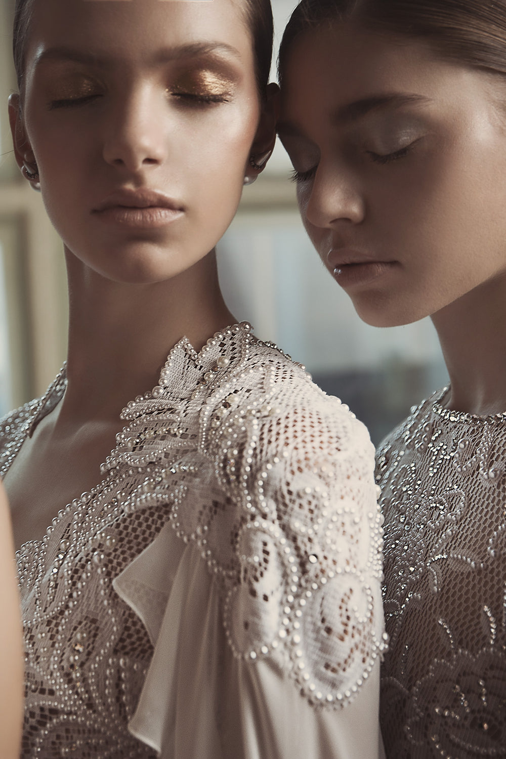 שתי נשים בשמלות כלה אלגנטיות ומורכבות, עומדות זו לצד זו עם מבט שלווה ועיניים עצומות, ומציגות את הפרטים העדינים המעוטרים בפנינים מקולקציית הכלות של ג'ולי לשנת 2018.