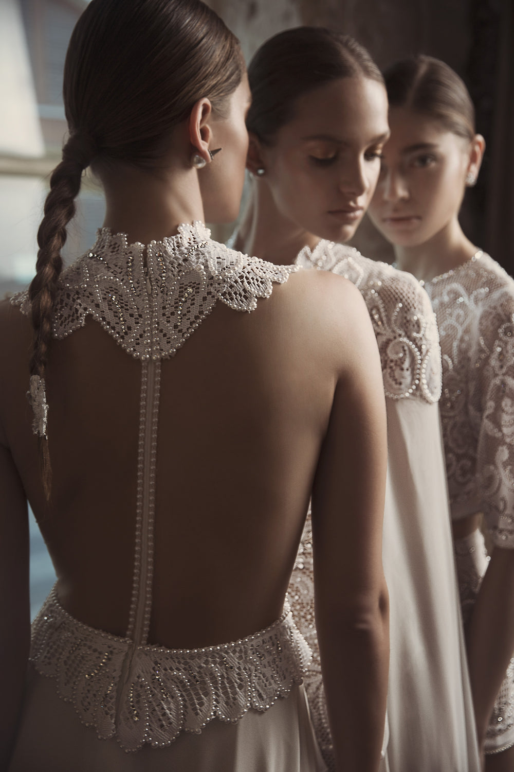 שלוש נשים בשמלות כלה אלגנטיות מקולקציית הכלות 2018 - Julie's by דרור קונטנטו עומדות זו לצד זו. המוקד הוא על רקמת חרוזים ותחרה מורכבת, כאשר שתי הנשים האחרות מטושטשות קלות ברקע.