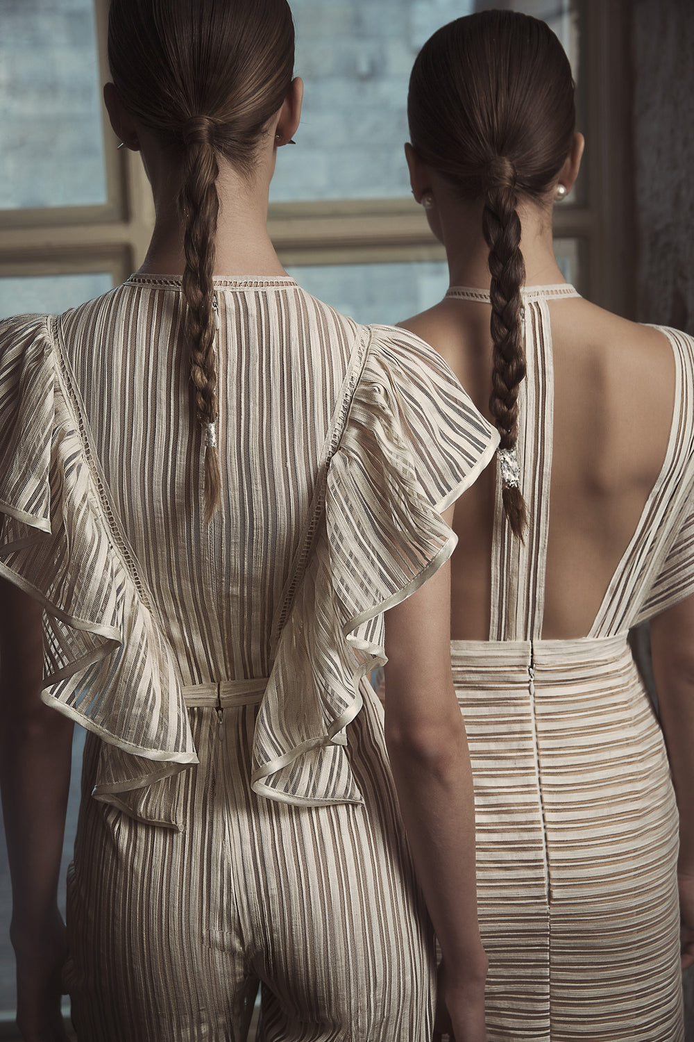 שתי נשים עם צמות ארוכות עומדות זו לצד זו, מדגמנות את קולקציית הכלות 2018 של דרור קונטנטו - טיאנה + יארה. הן לובשות שמלות כלה תואמות בצבע שמנת ובז' עם שרוולים מקושטים וגב פתוח, מוארות באור טבעי רך הנכנס מהחלון.