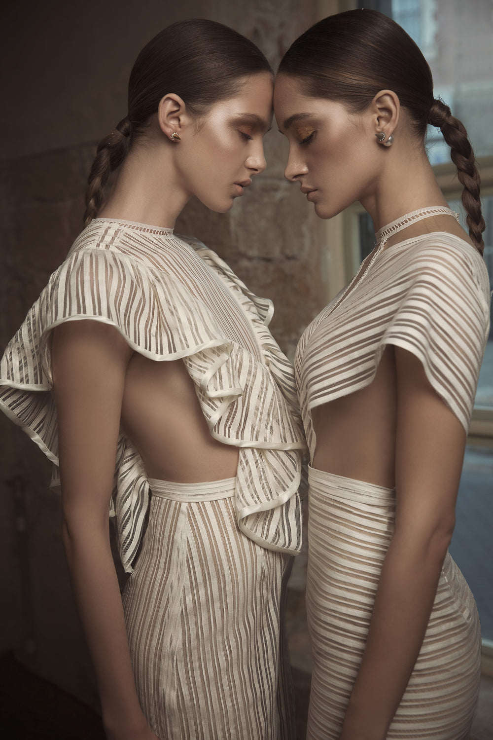 שתי נשים עם שיער קלוע עומדות זו מול זו, עיניהן עצומות, בחדר מואר באור רך, לבושות בשמלות תואמות עם פסים קרמיים ורופפות מקולקציית הכלות של דרור קונטנטו 2018 - טיאנה + יארה.