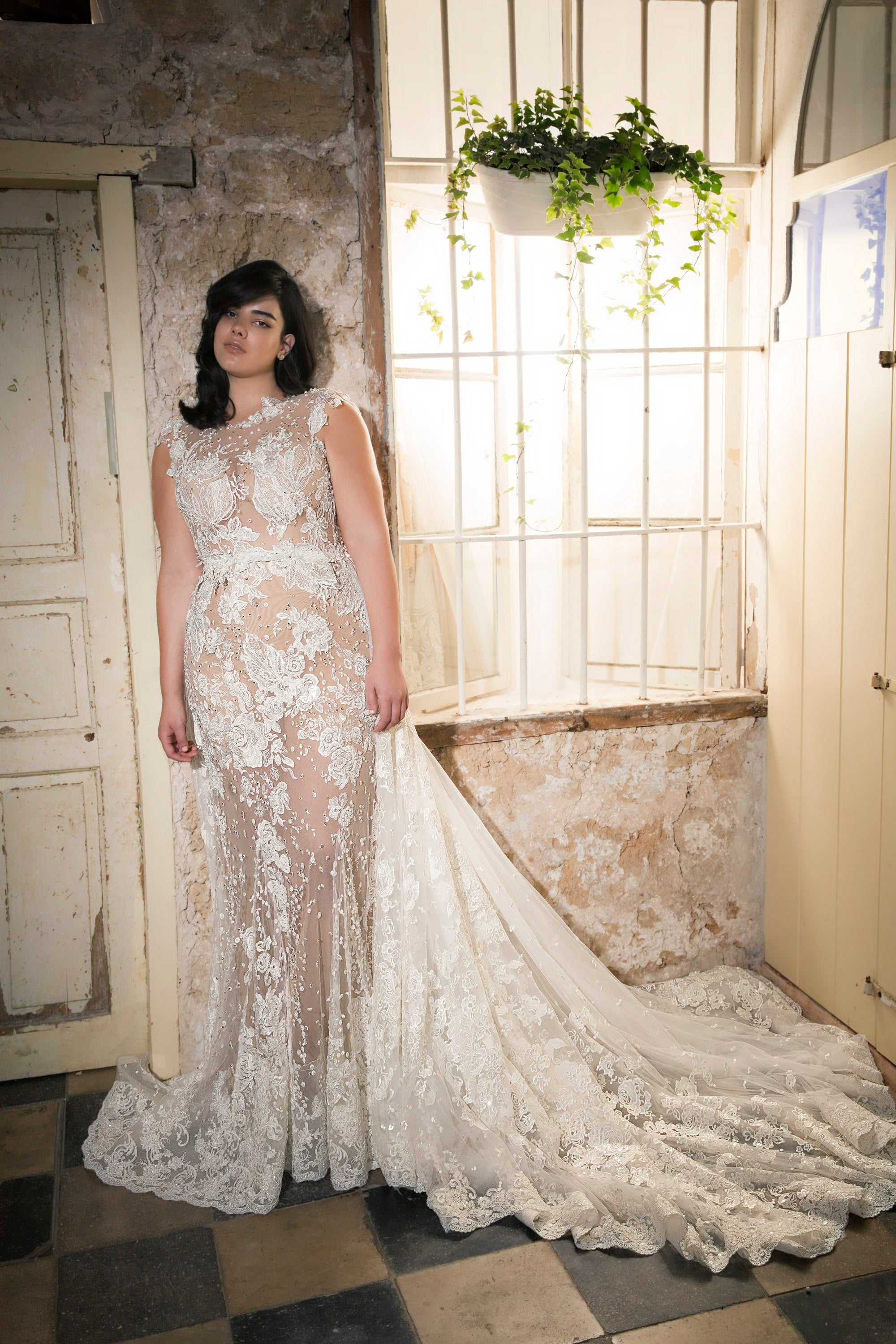 אישה לבושה בשמלת כלה מקולקציית Bridal Plus 2017 - Simone עומדת בתוך הבית ליד חלונות גדולים עם צמחים משתלשלים, על רקע כפרי של אבן ועץ.